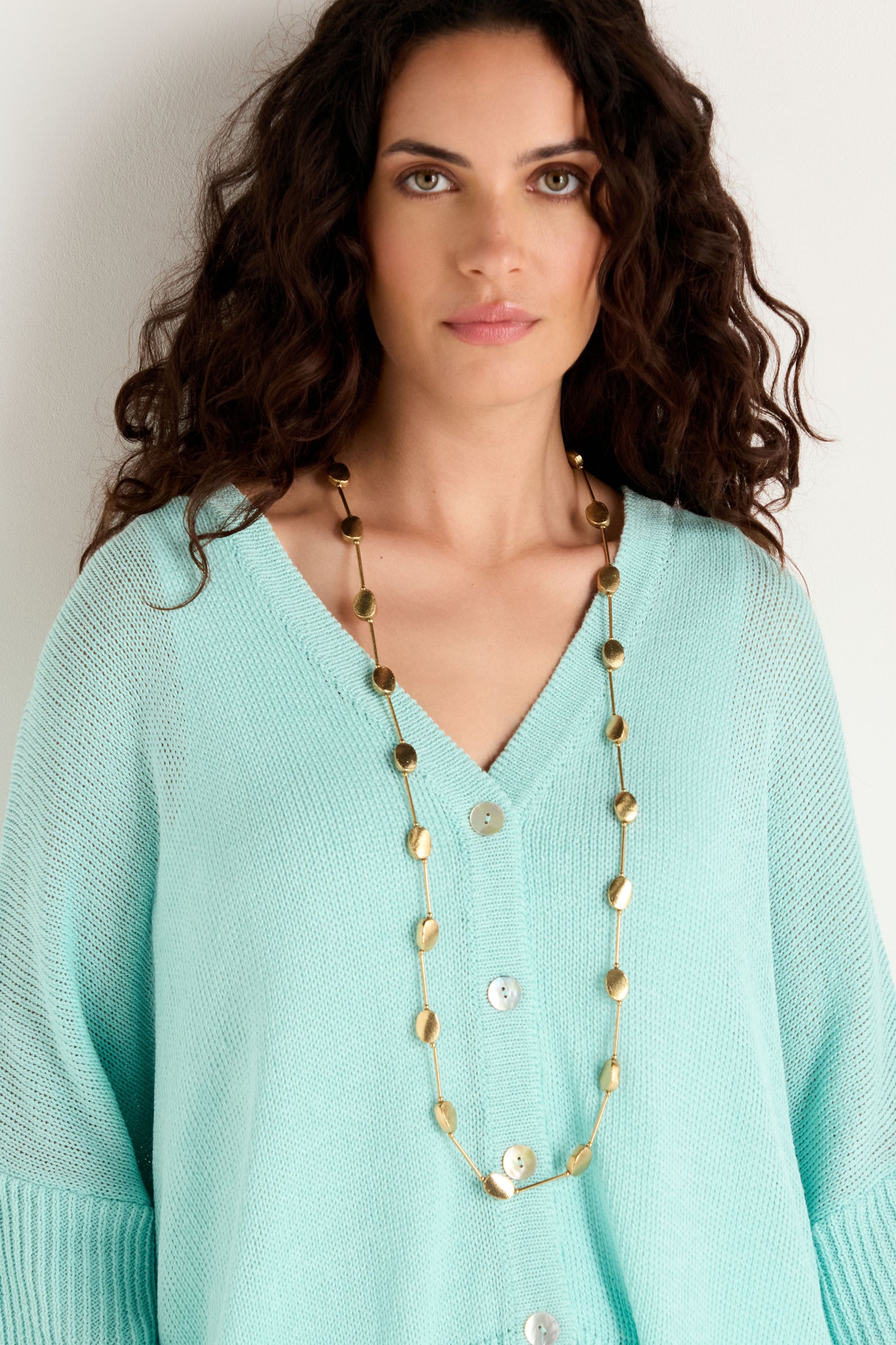 A woman stands against a plain light background, wearing a light blue buttoned sweater and the Long Oval Disc Necklace featuring textured oval discs.