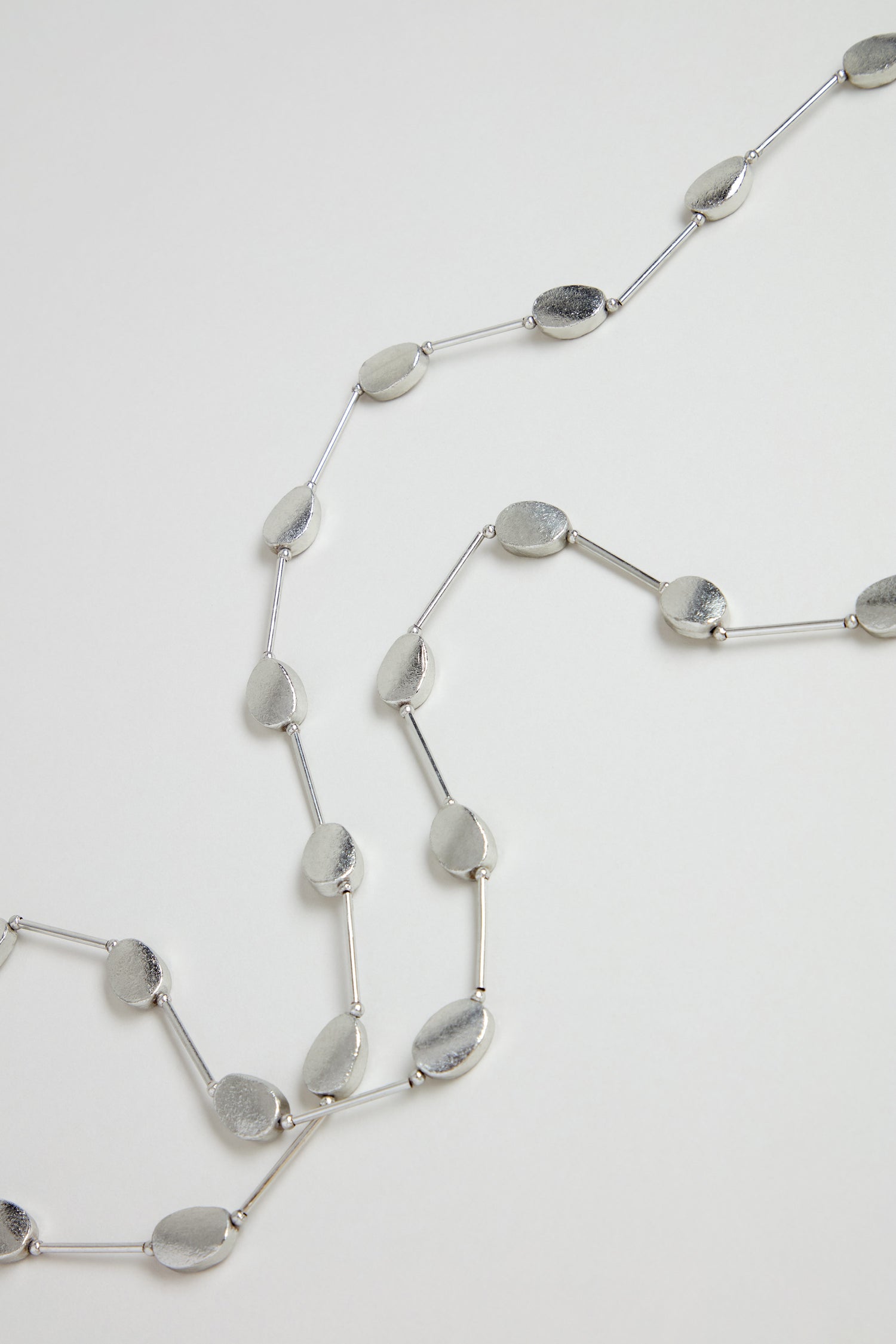 The Long Oval Disc Necklace showcases textured oval discs and slim connecting bars, elegantly displayed on a white background.
