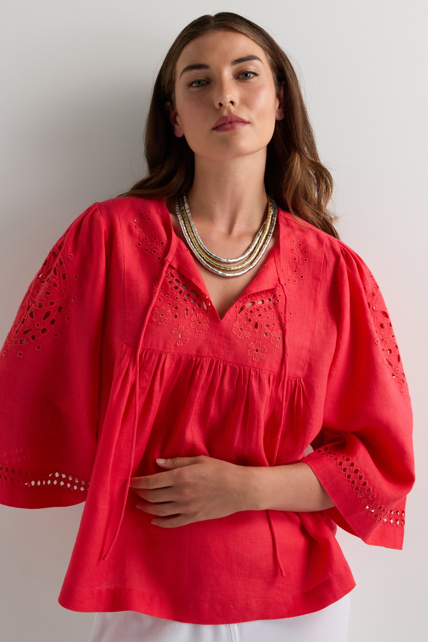 A woman in a red blouse with eyelet sleeve details wears the Triple Strand Disc Necklace, which features metallic-toned discs, as she stands against a light background.