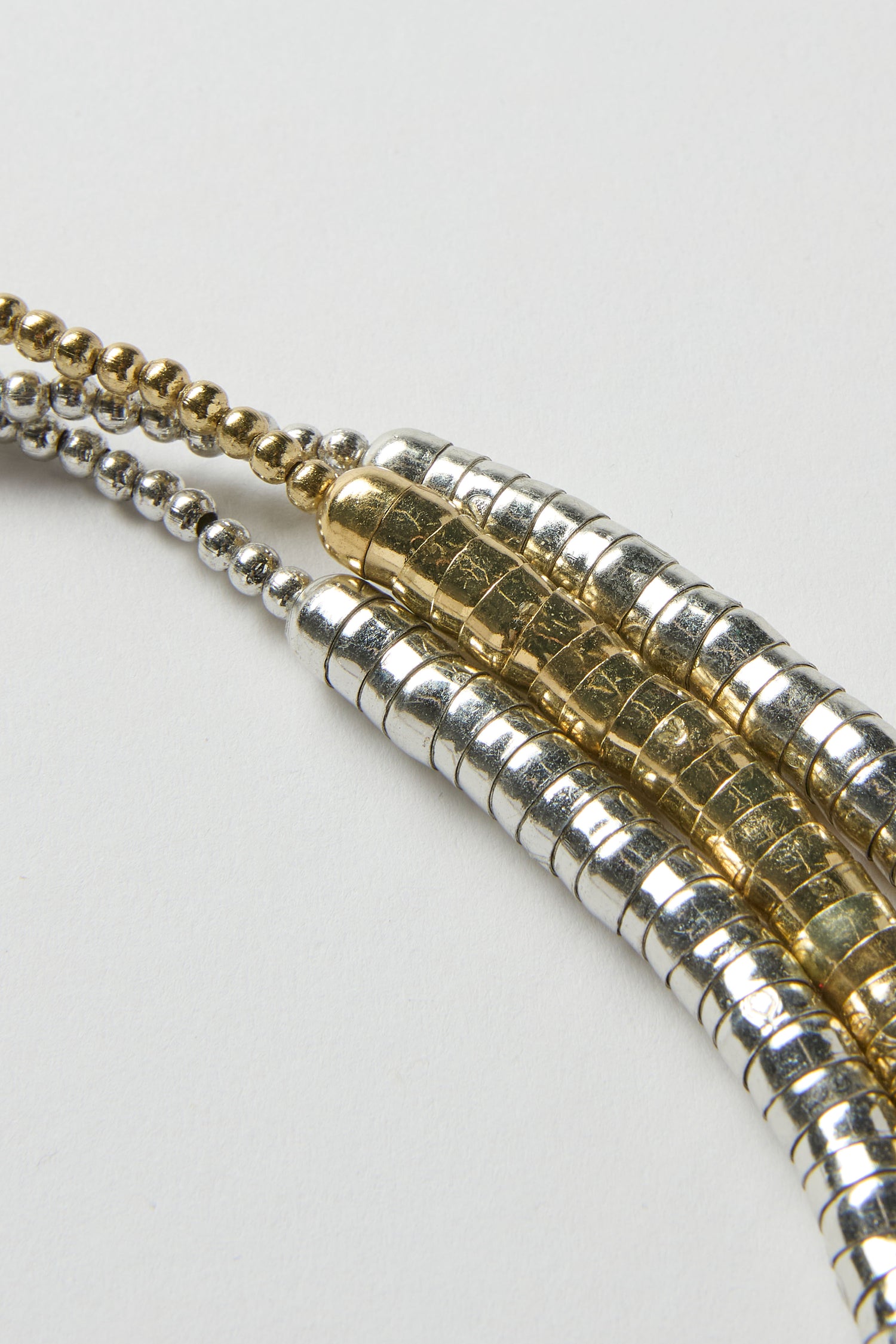 Close-up of the Triple Strand Disc Necklace, featuring three parallel metallic beaded chains—two silver and one gold—arranged on a white surface.