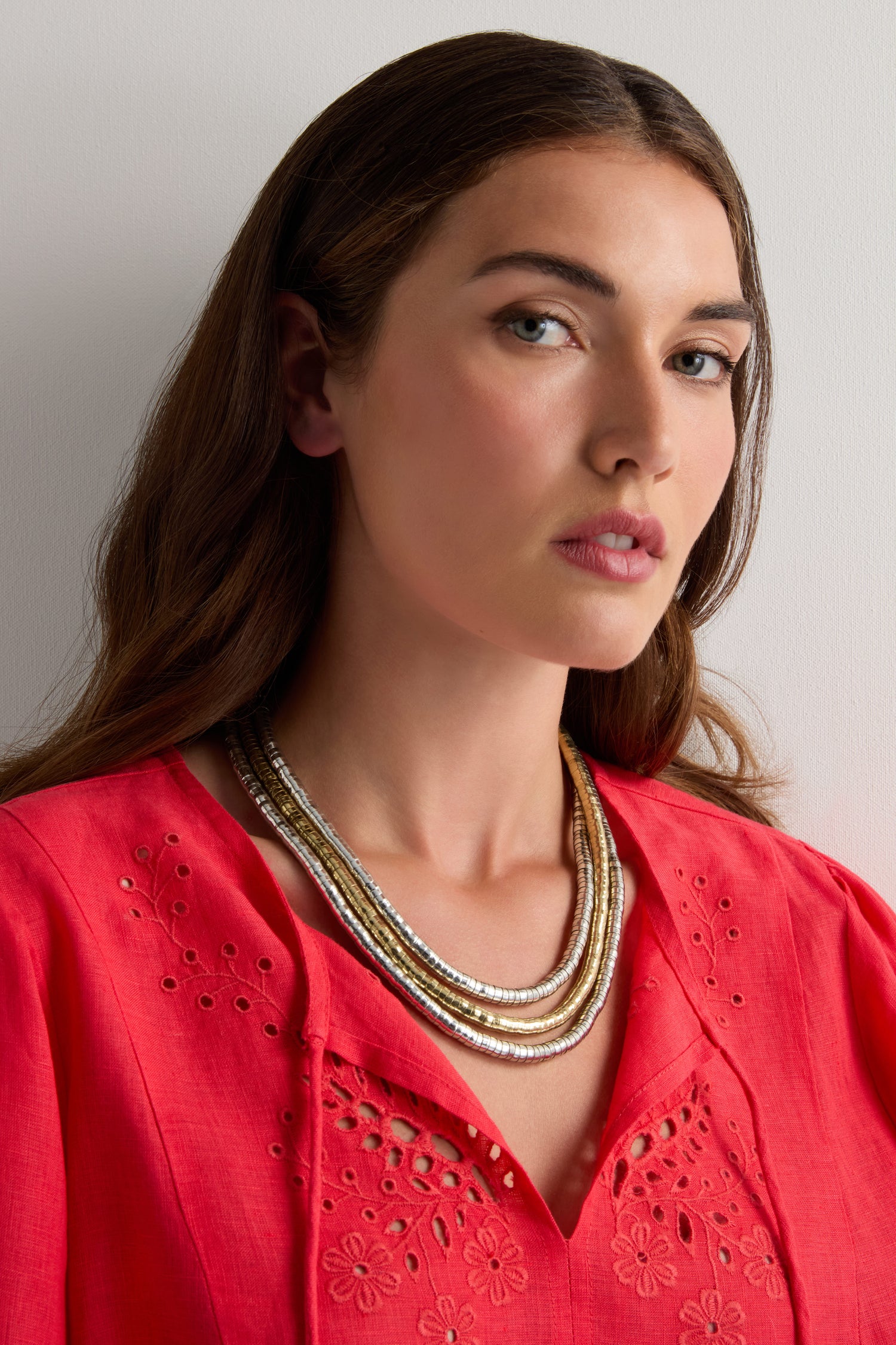 A woman with long brown hair models a coral eyelet-embroidered blouse and the Triple Strand Disc Necklace, featuring layered metallic-toned discs, against a plain light background.