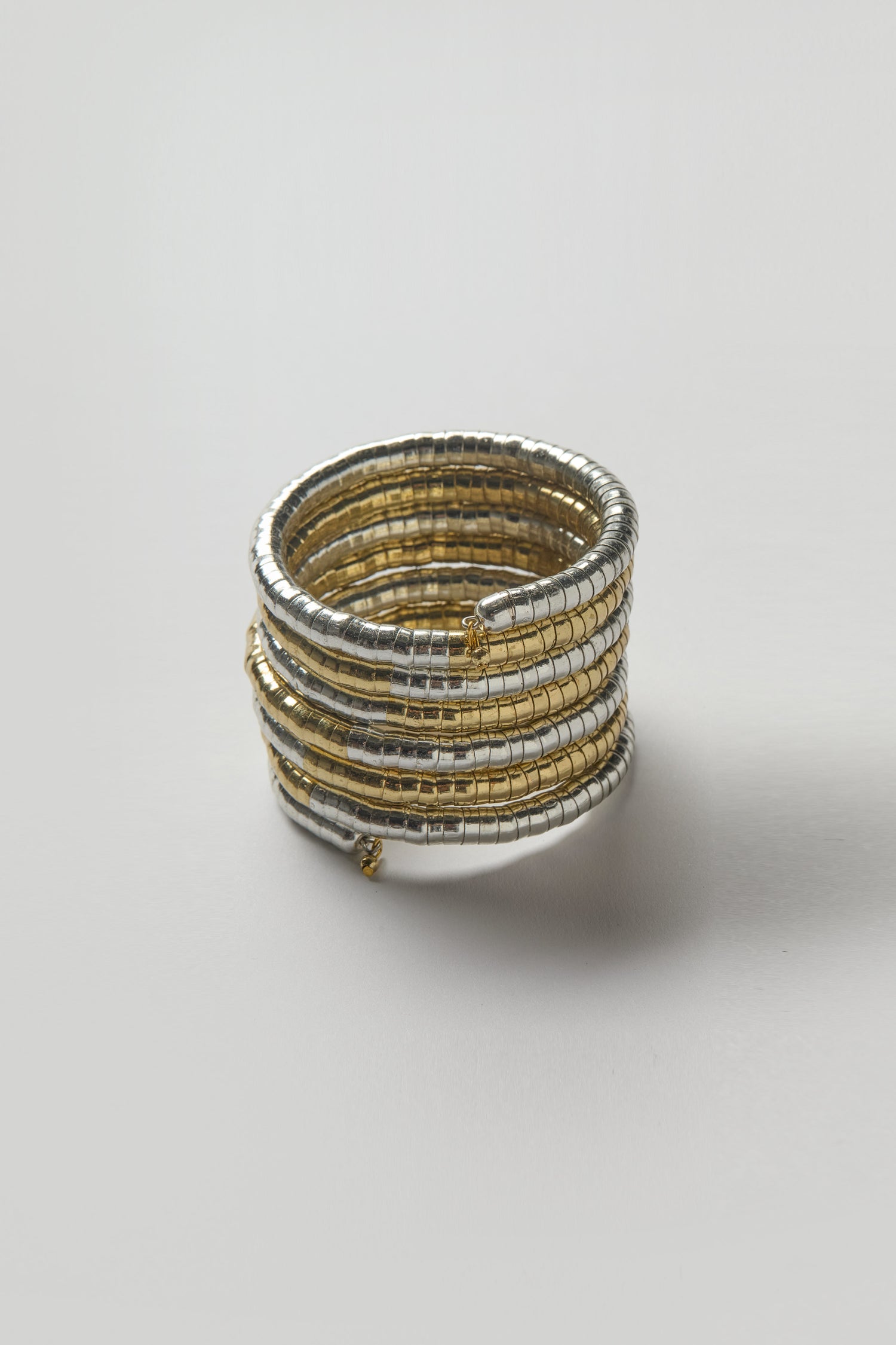 The Mixed Metal Spiral Cuff showcases alternating gold and silver tones, creating a sculptural, coiled silhouette against a plain white background.