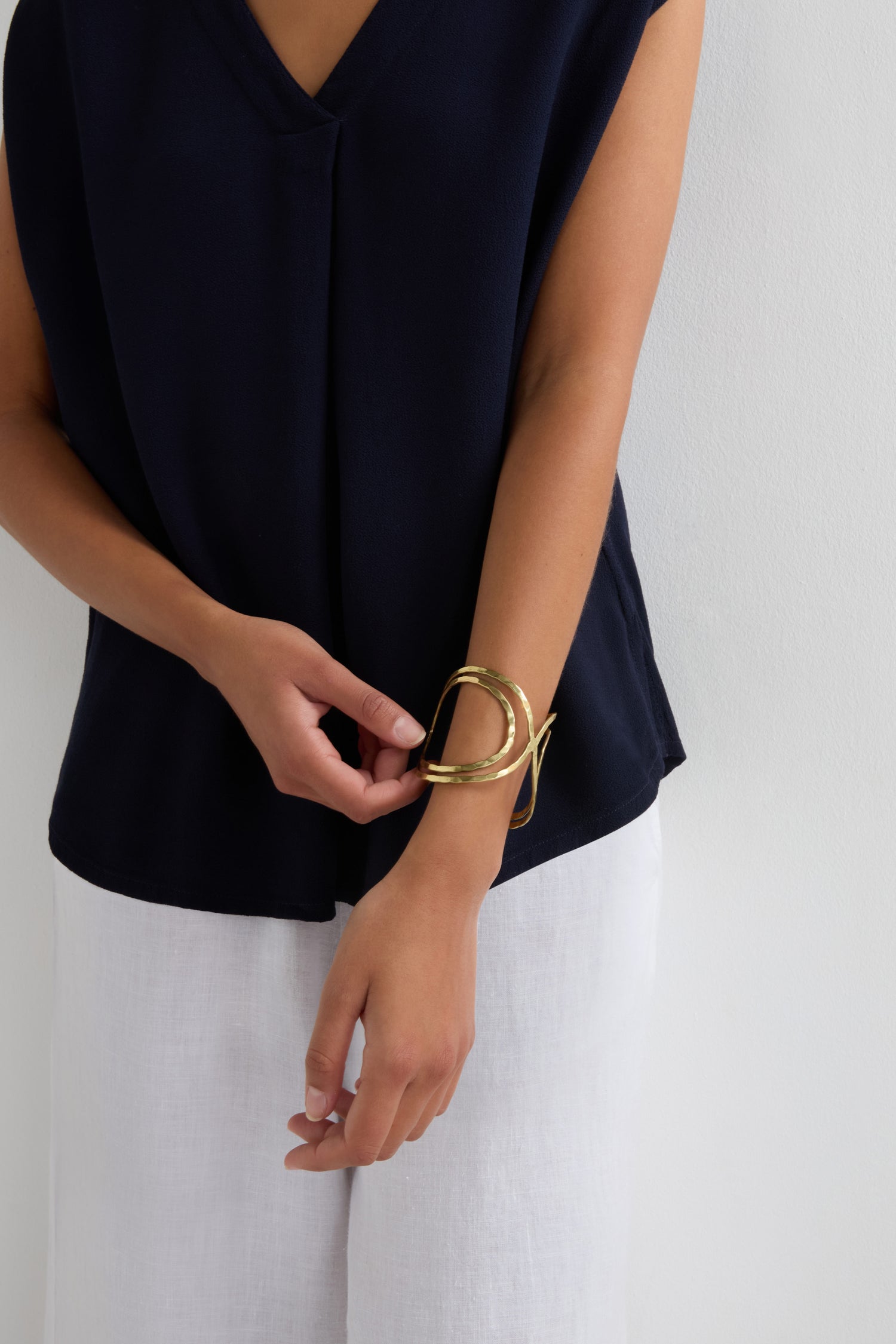 A person wearing a navy sleeveless top and white pants adjusts the Butterfly Bangle, a large gold cuff-style bracelet, on their wrist.
