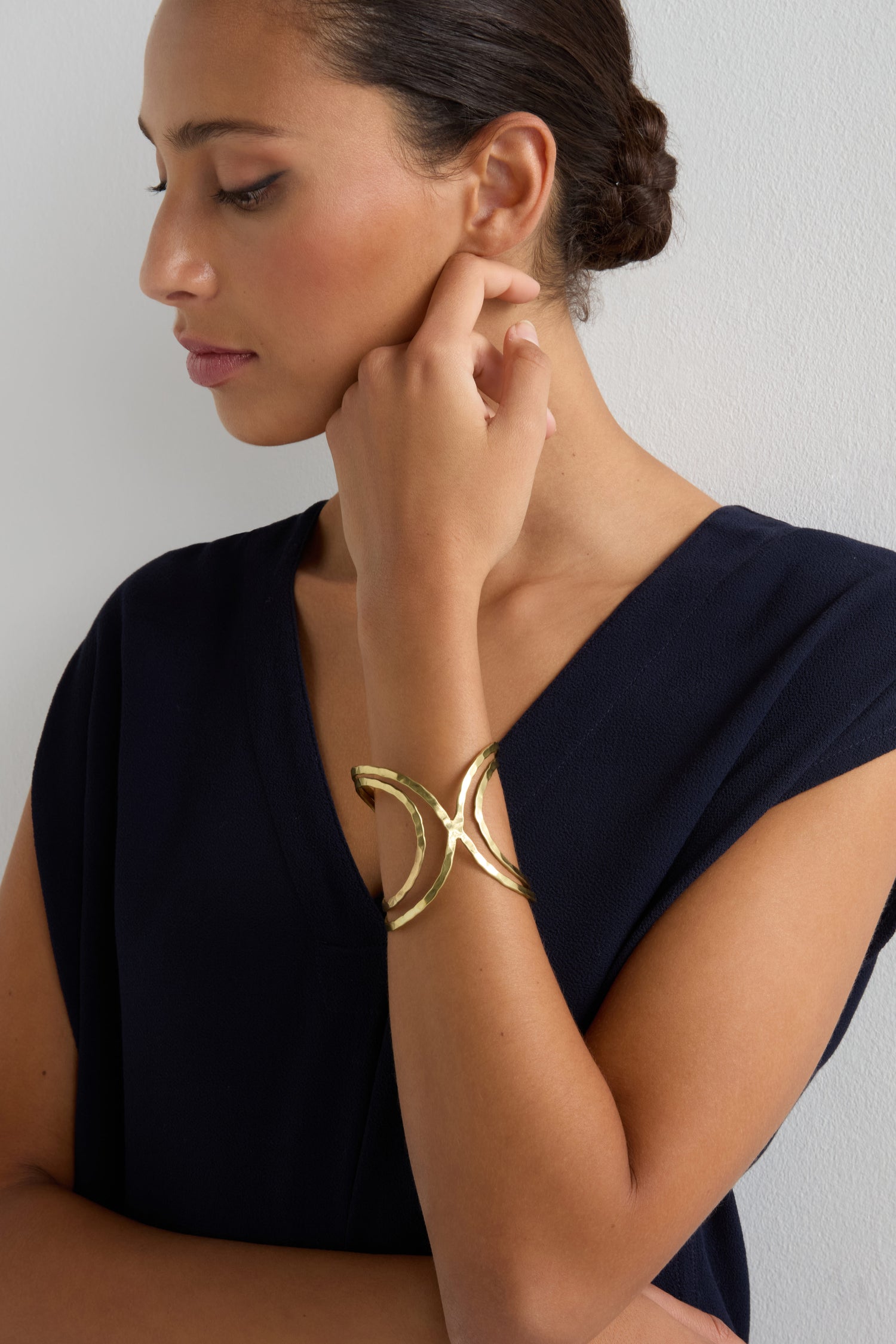 A woman in a navy V-neck top wears the Butterfly Bangle—a large, gold cuff-style bracelet with a sculptural silhouette—on her wrist, her hair pulled back and hand raised to her chin.