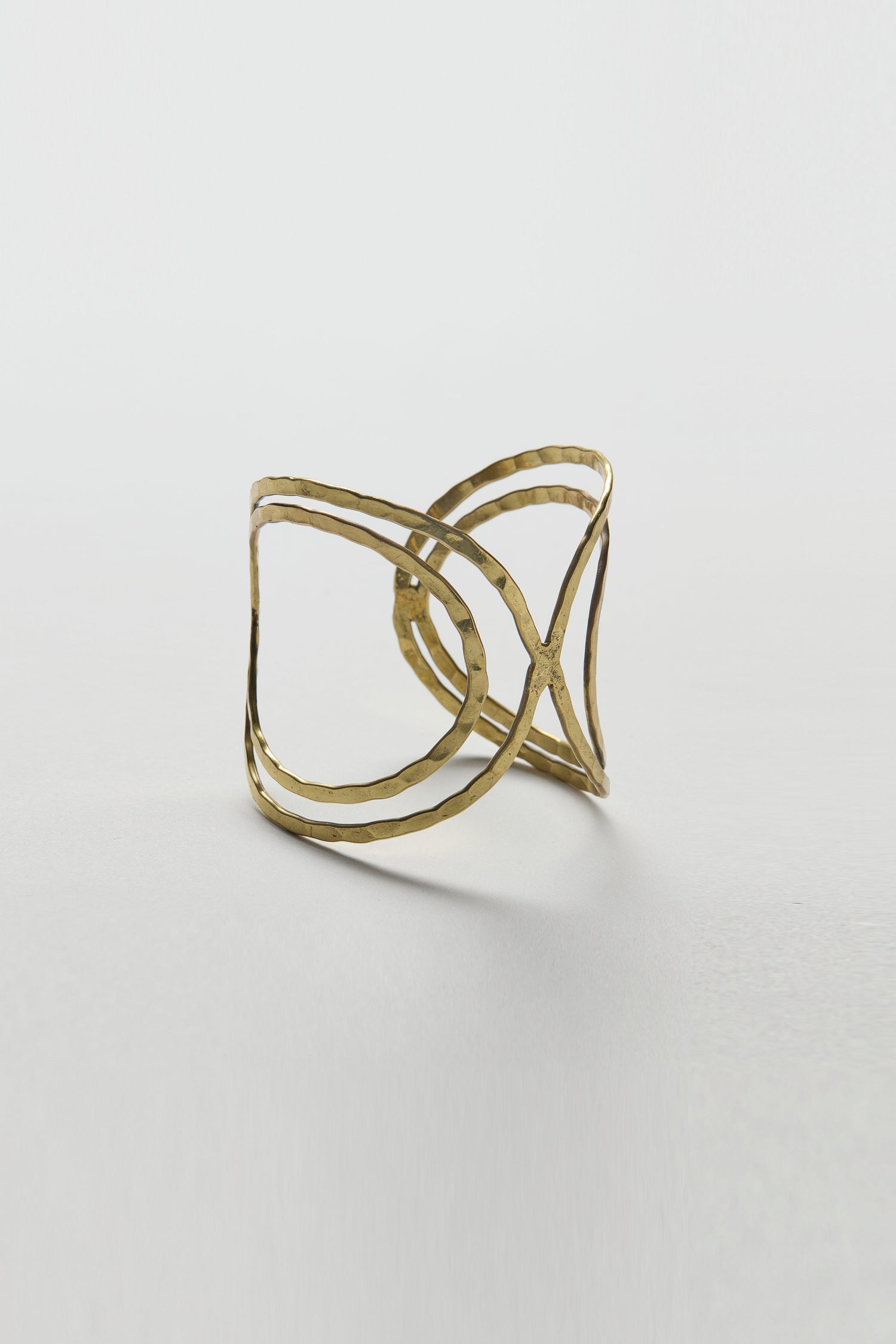 The Butterfly Bangle is a gold cuff-style bracelet with a hammered texture, featuring an intersecting crisscross design and sculptural silhouette, displayed on a plain white background.