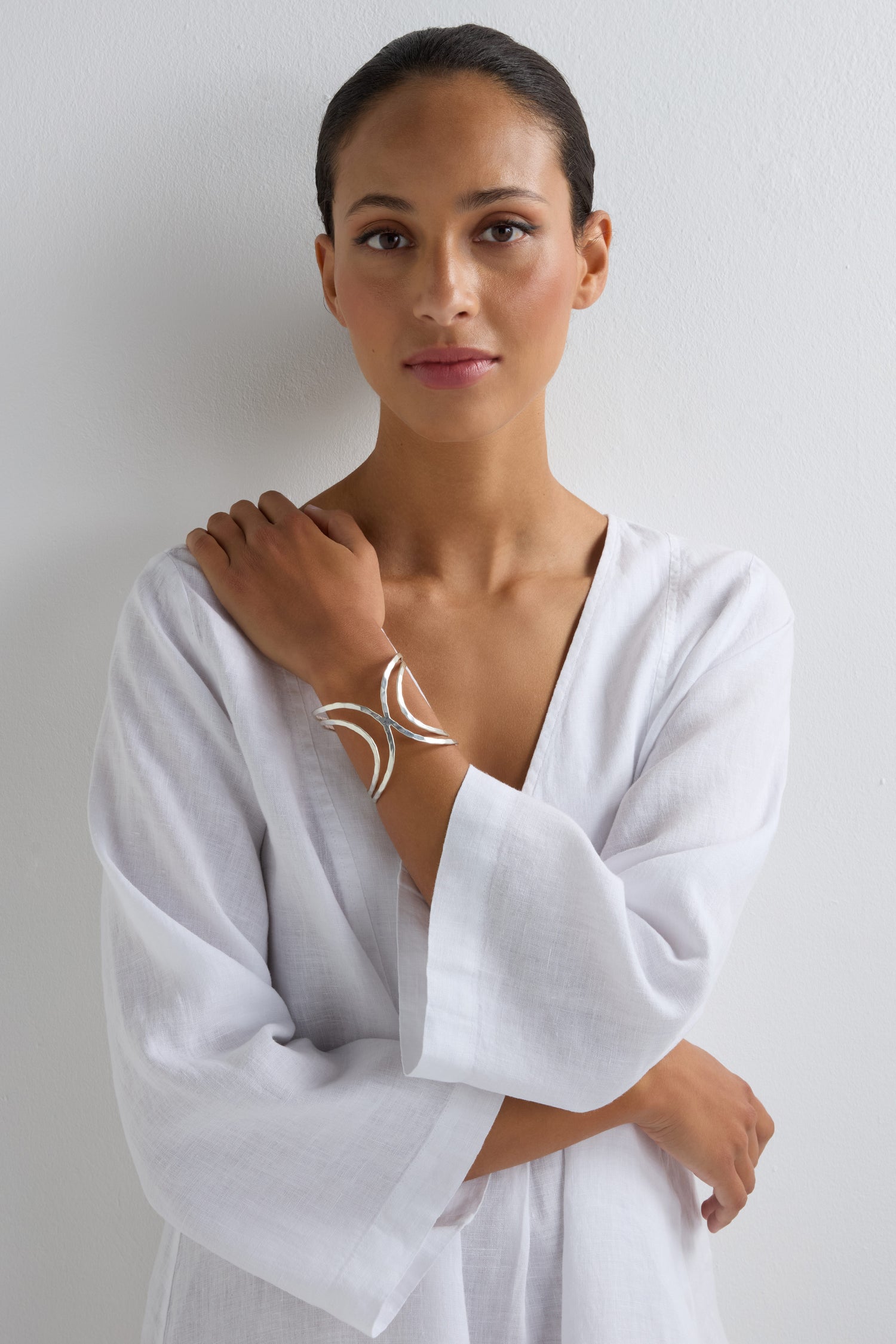 A woman in a white, loose-fitting garment stands against a white wall, wearing the modern Butterfly Bangle on her left wrist and looking directly at the camera.