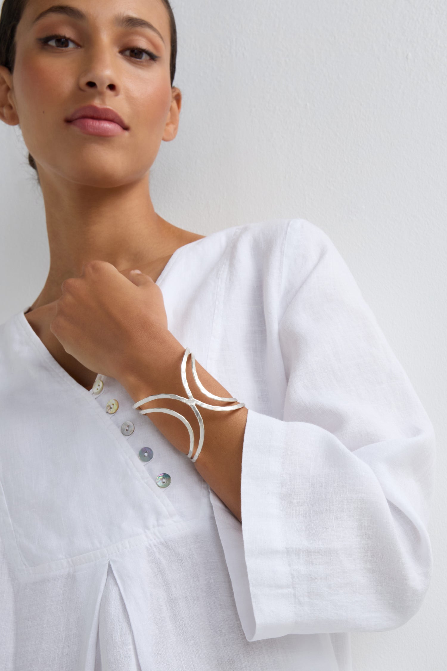 A woman poses against a plain white background, wearing a white blouse with button details and the Butterfly Bangle—a large, silver cuff bracelet with a sculptural silhouette.