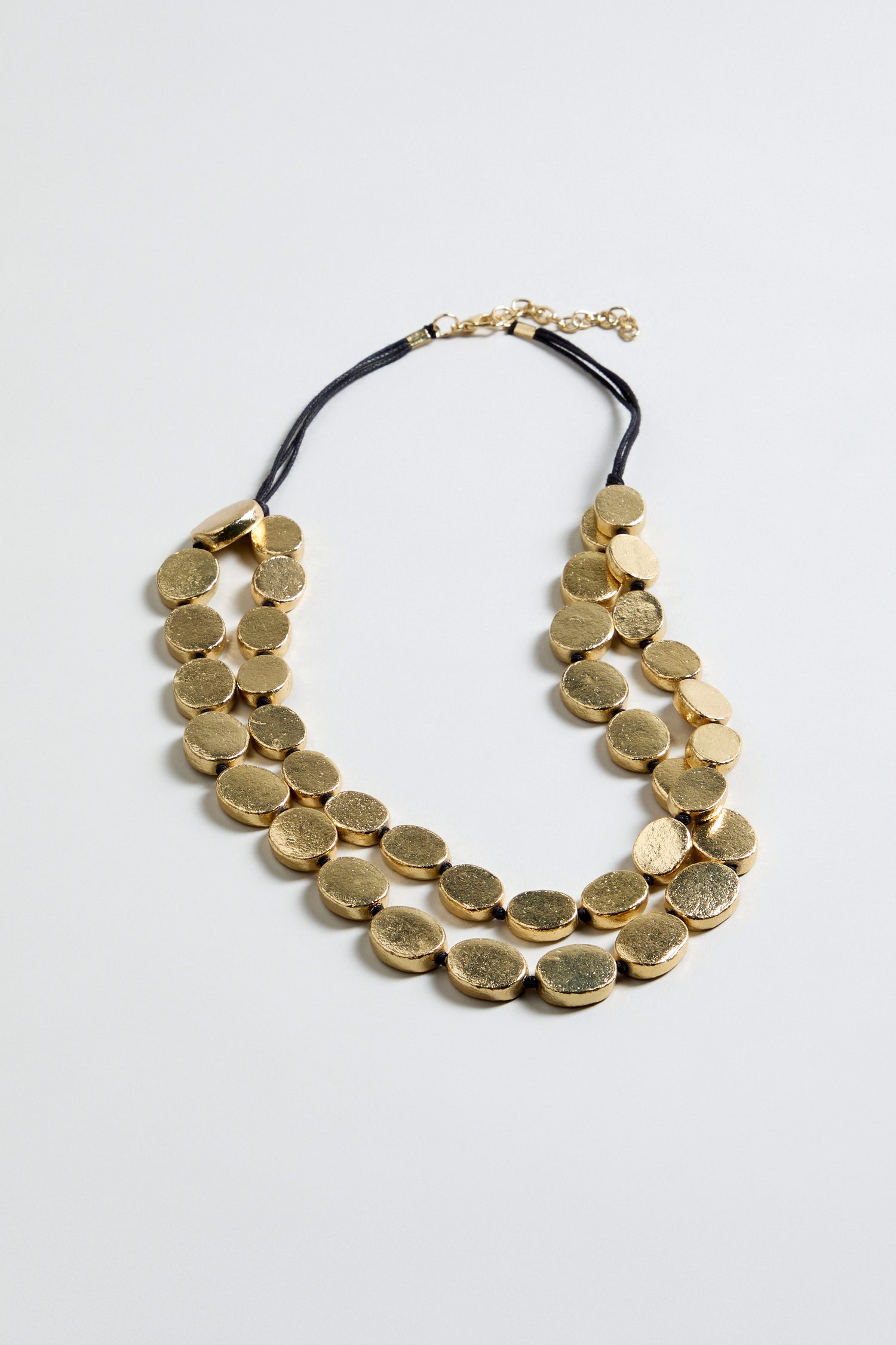 The Double Strand Oval Disc Necklace features two rows of gold-colored, textured oval discs on a black cord with an adjustable clasp, displayed against a plain light background.