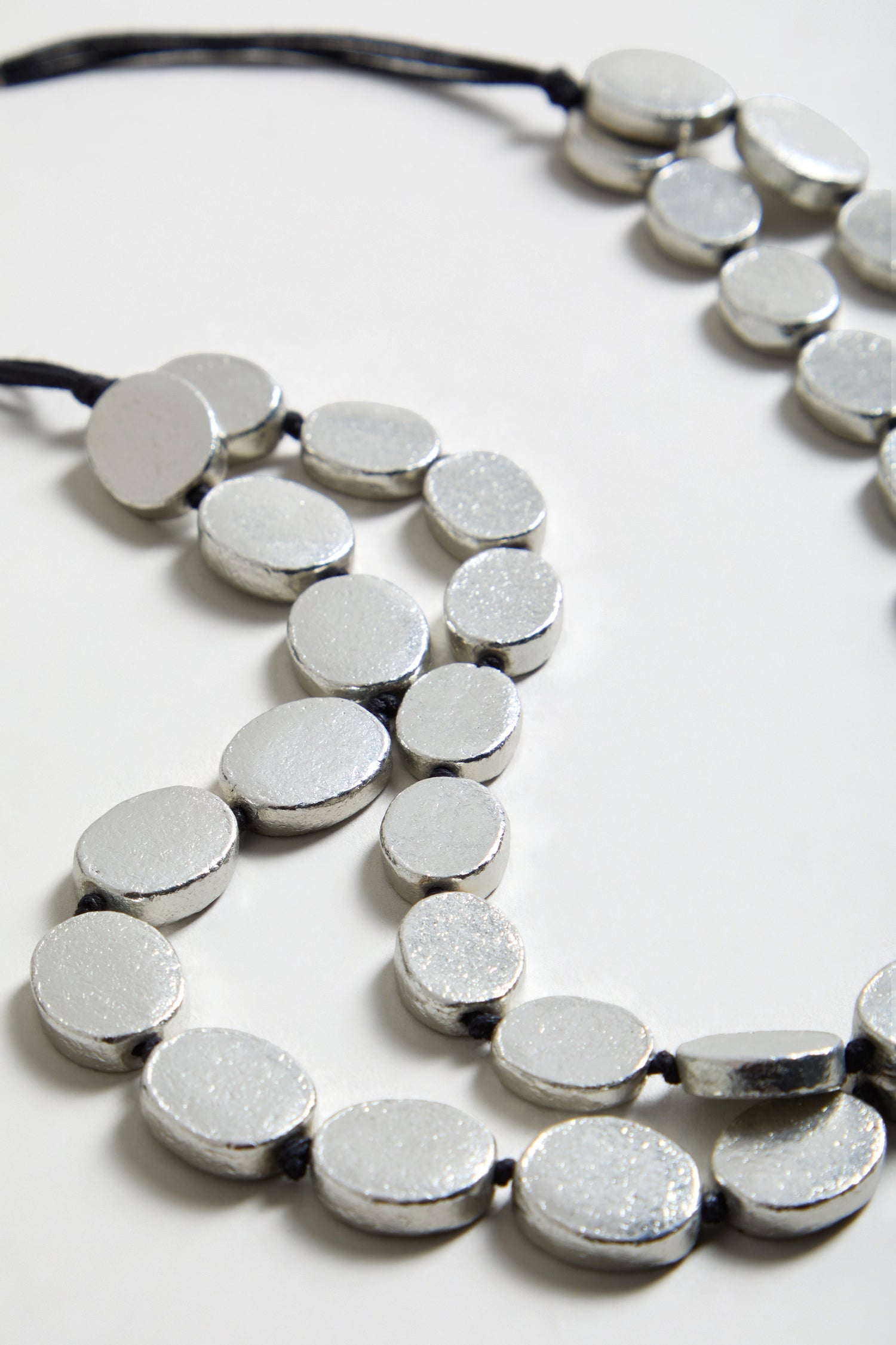 Close-up of two silver metallic beaded necklaces with black cords, including the Double Strand Oval Disc Necklace, arranged on a white surface.
