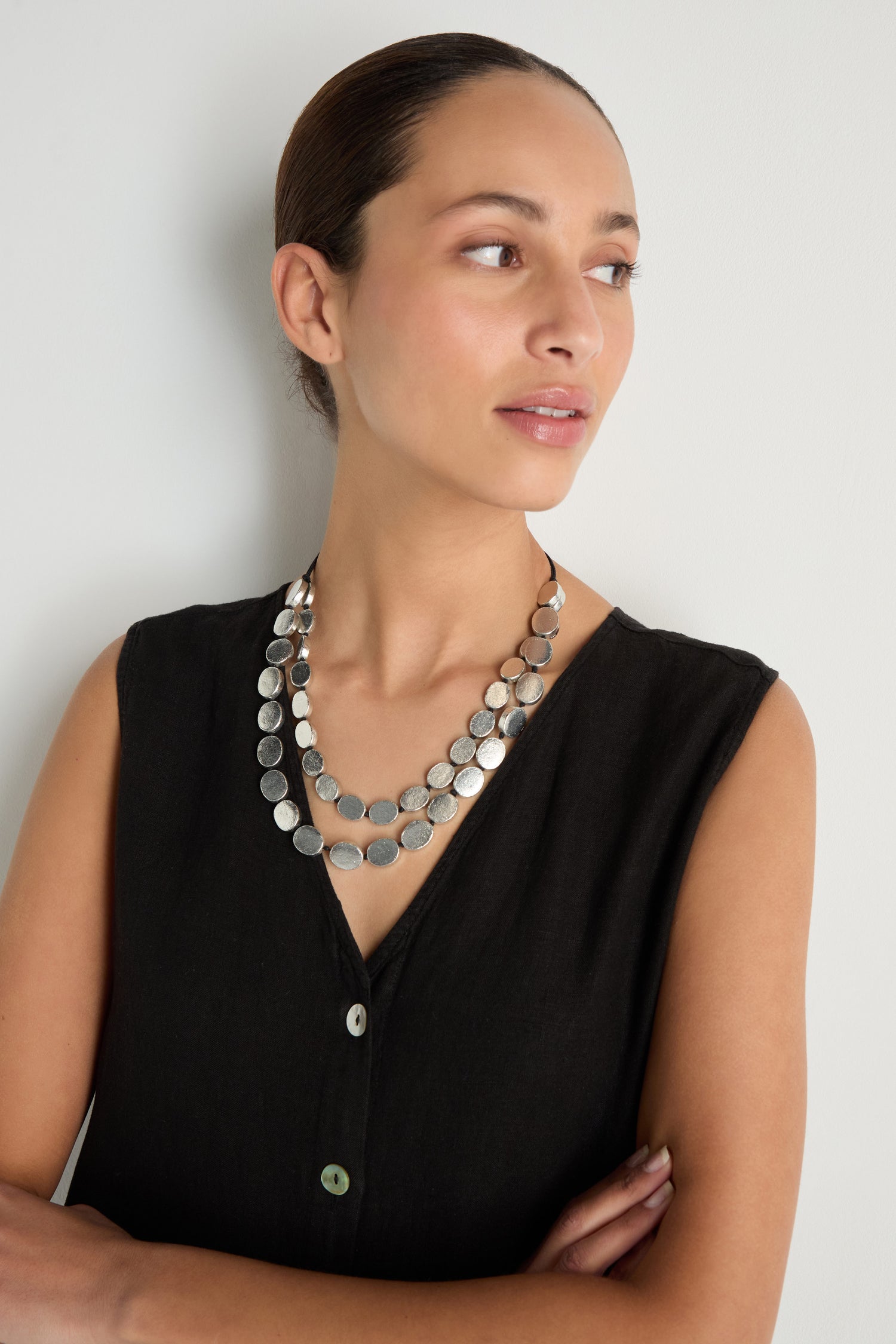 A woman in a sleeveless black top and the Double Strand Oval Disc Necklace with layered silver coin pendants stands against a white wall, arms crossed and looking to the side.