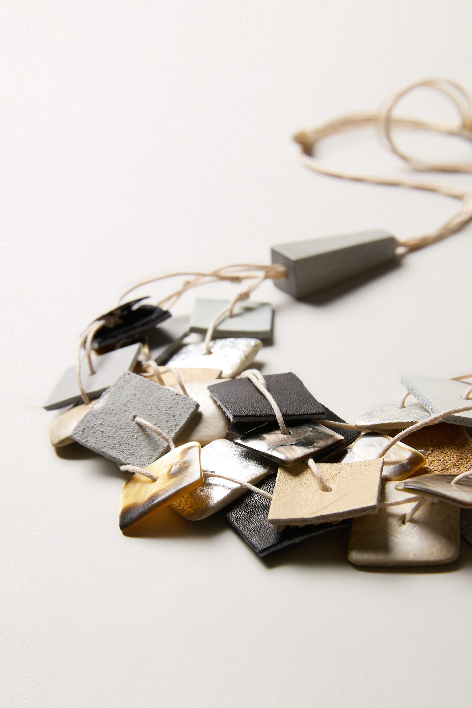 A close-up of the Gathered Shapes Necklace, showcasing geometric square and rectangular pendants with modern texture on a beige cord, set against a plain light background.