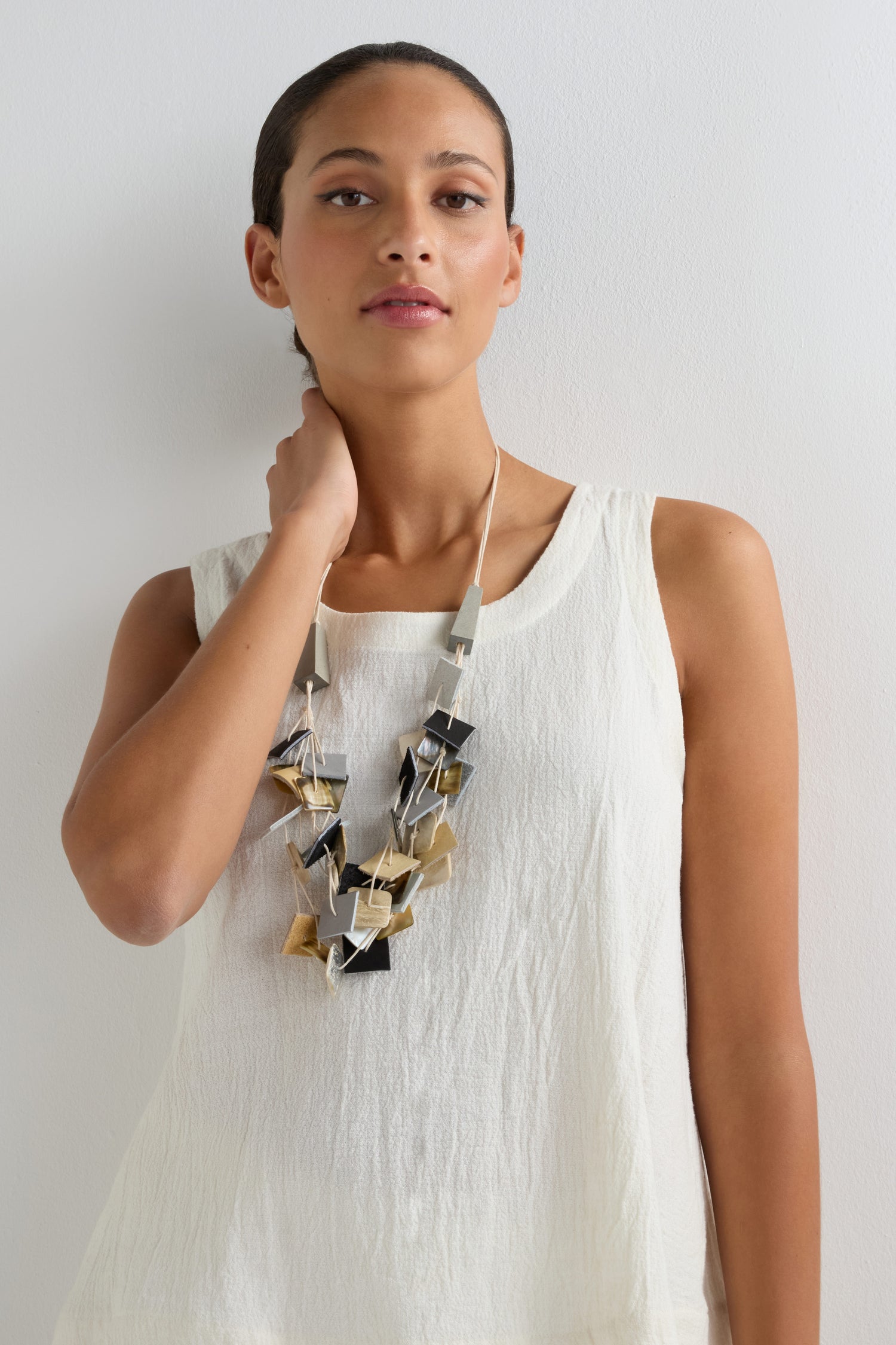 A woman in a sleeveless white top wears the Gathered Shapes Necklace, known for its bold geometric forms, and stands against a plain white background with one hand touching her neck.