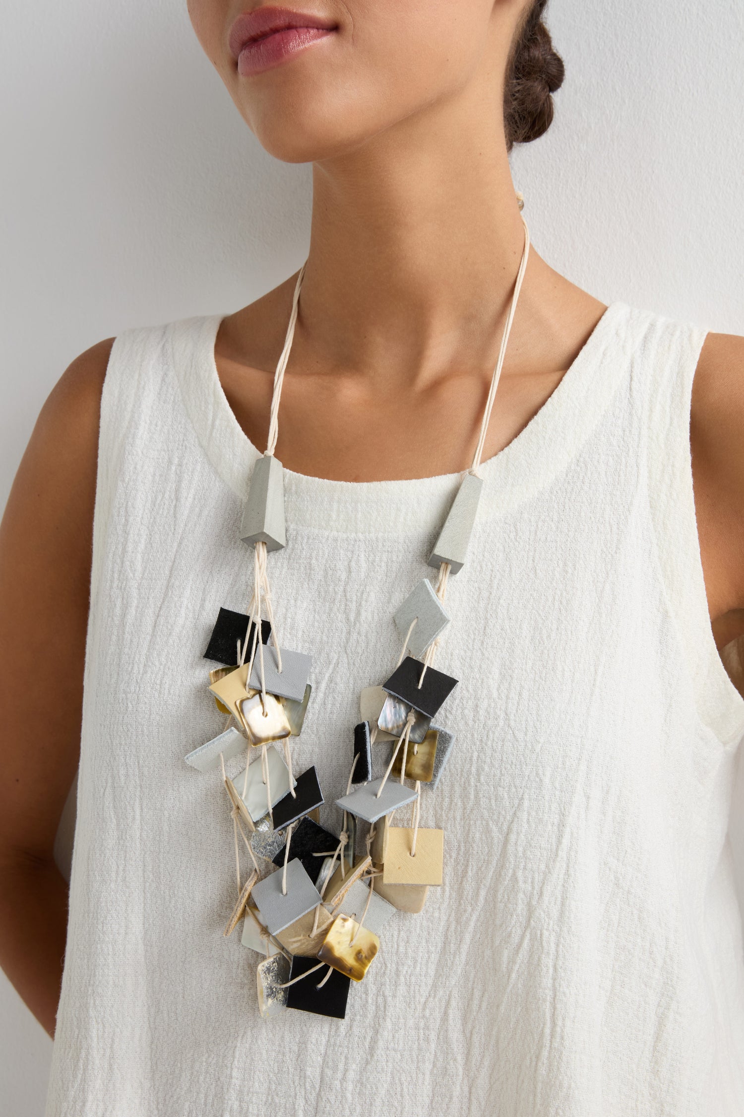 A woman wears a white sleeveless top paired with the Gathered Shapes Necklace, featuring geometric forms in gray, gold, and black tones.