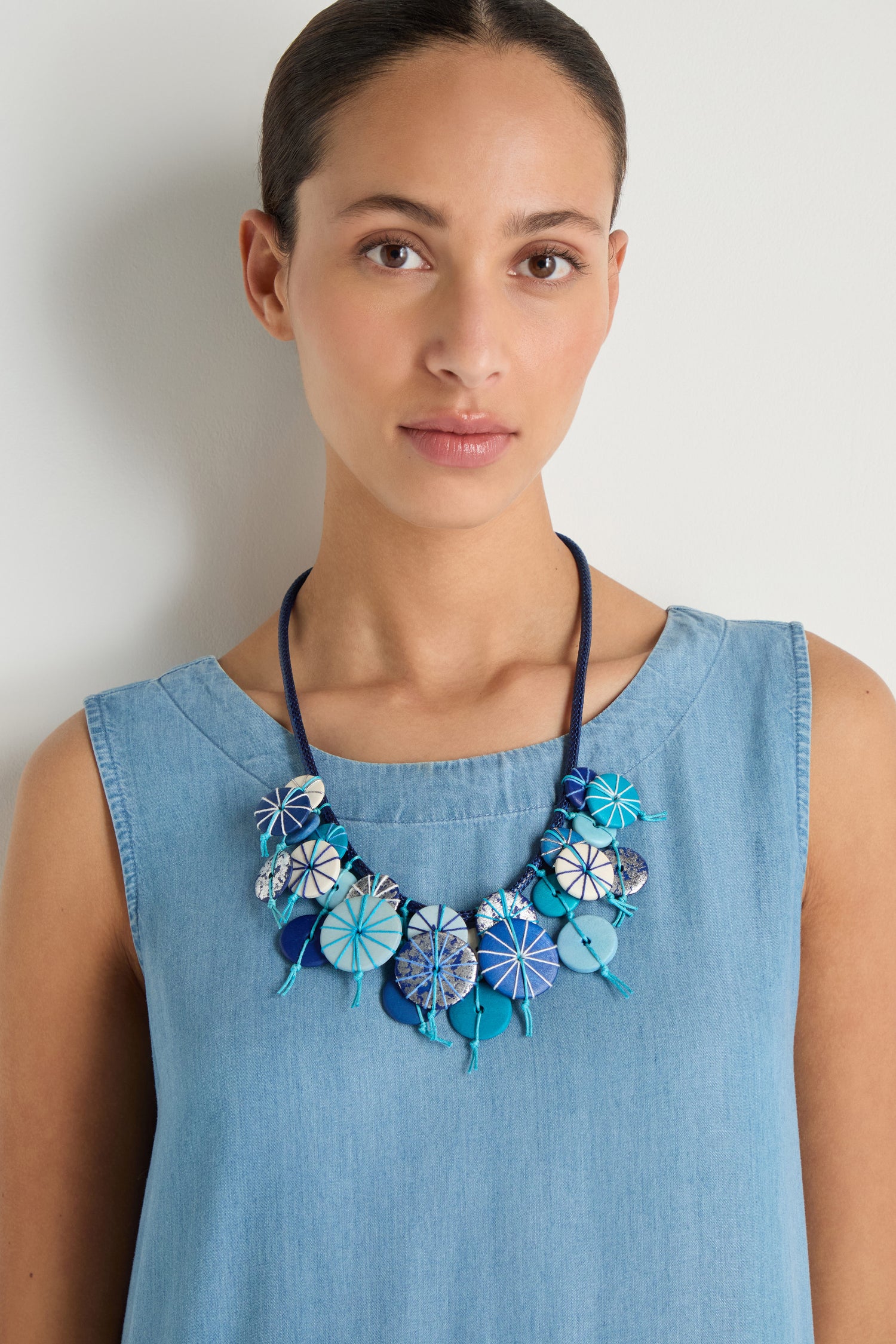 A woman in a sleeveless light blue dress wears the Azure Short Disc Necklace, featuring striking patterned circular elements, as she stands against a plain light background.