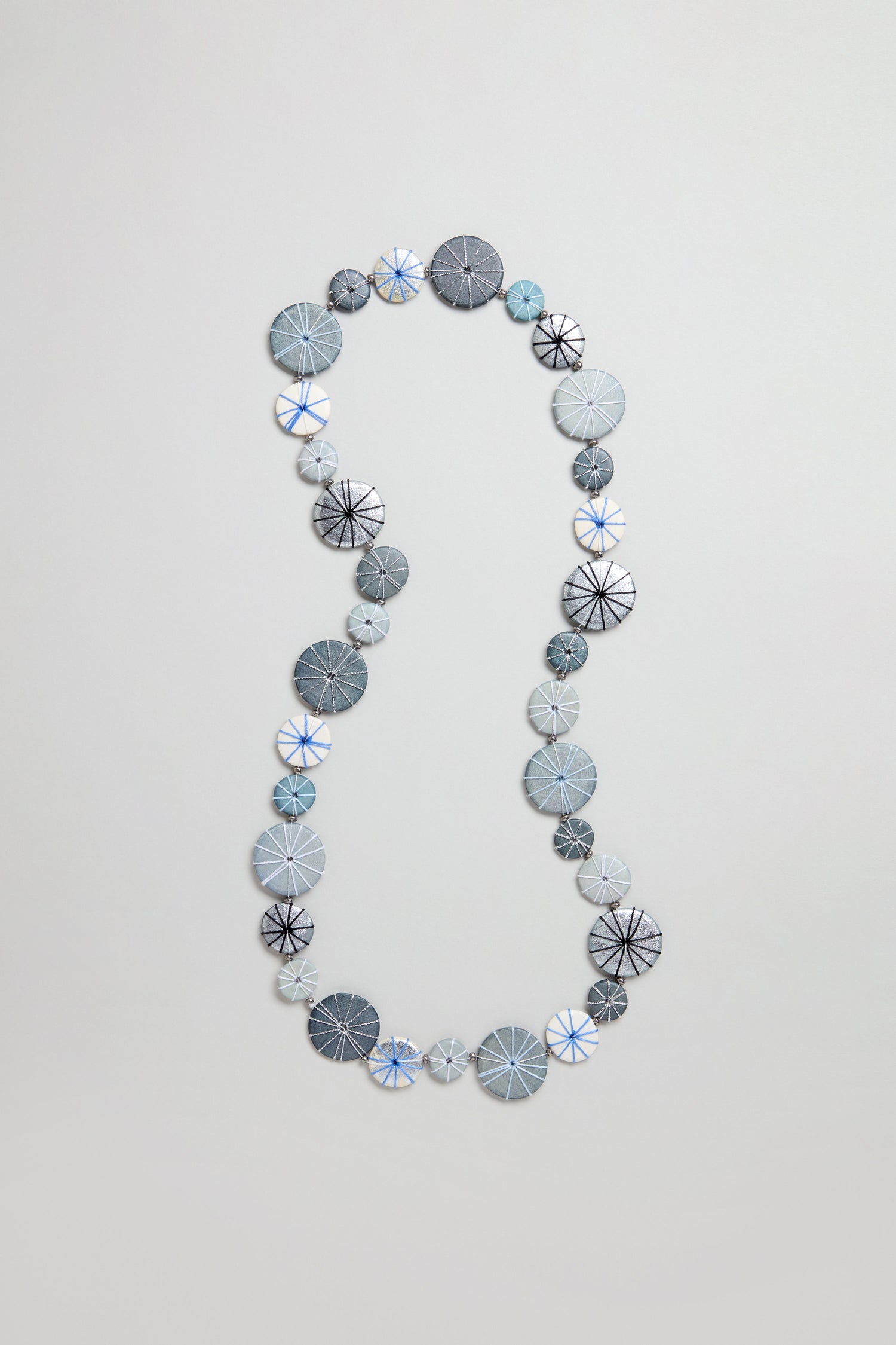 The Long Wooden Metallic Parasols Necklace features round, flat beads in gray, blue, and white with line patterns and subtle cotton accents, arranged in a loose oval on a plain background.