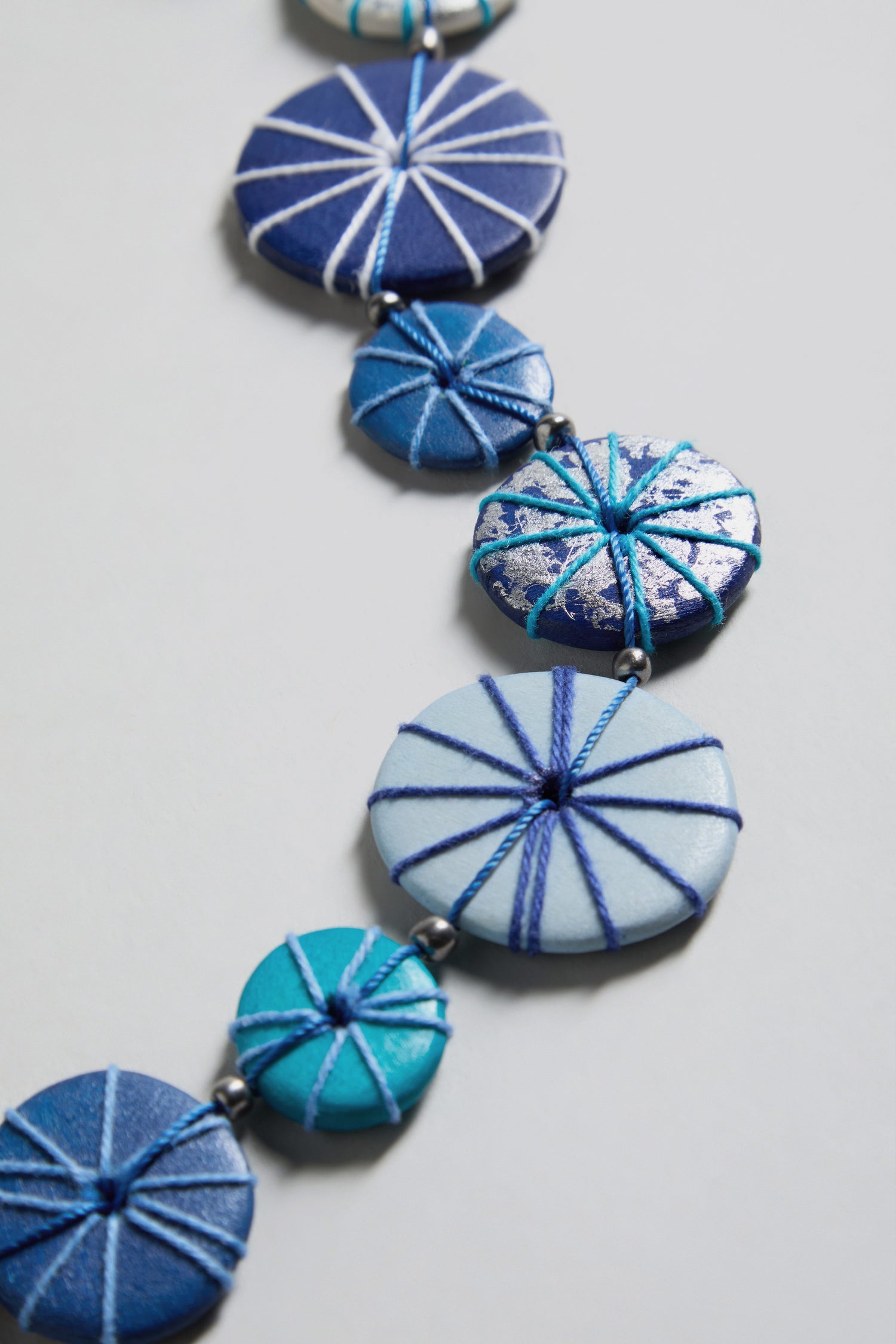 The Long Wooden Disc Necklace features flat wooden beads in various blue hues with detailed blue and white thread wraps, displayed on a light surface.