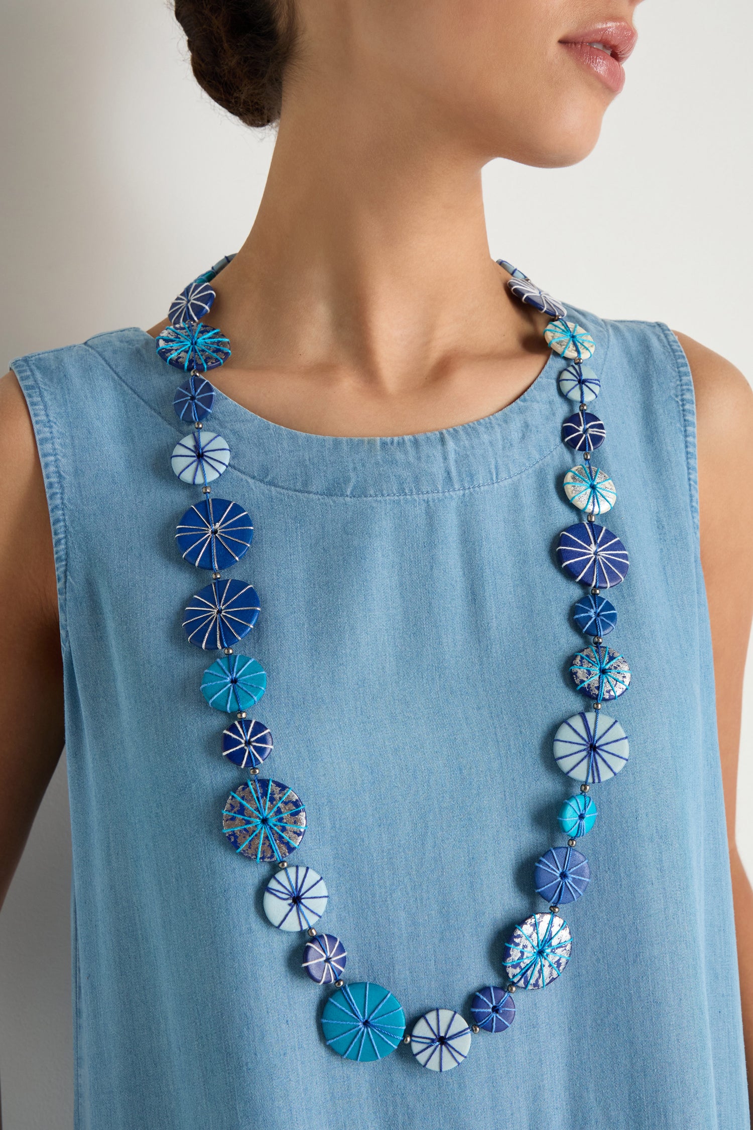 A woman in a sleeveless light blue dress showcases the Long Wooden Disc Necklace, which features blue and white patterned beads, an adjustable cord, and thread wrap detail.