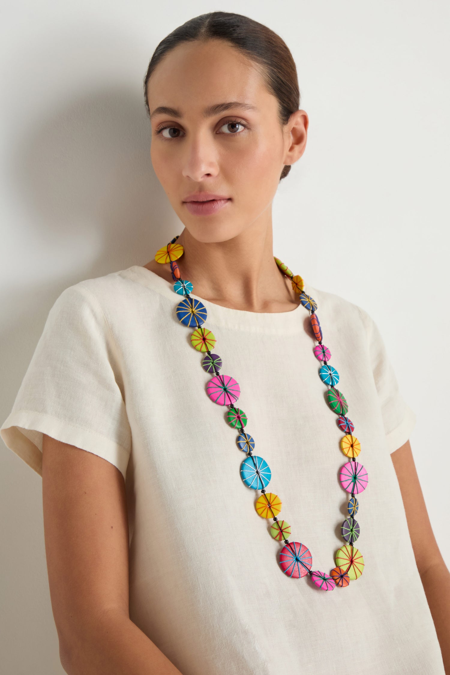 A woman stands against a plain light background wearing a cream short-sleeve top and the Long Wooden Disc Necklace—an adjustable cord necklace with circular, multicolored wooden beads.