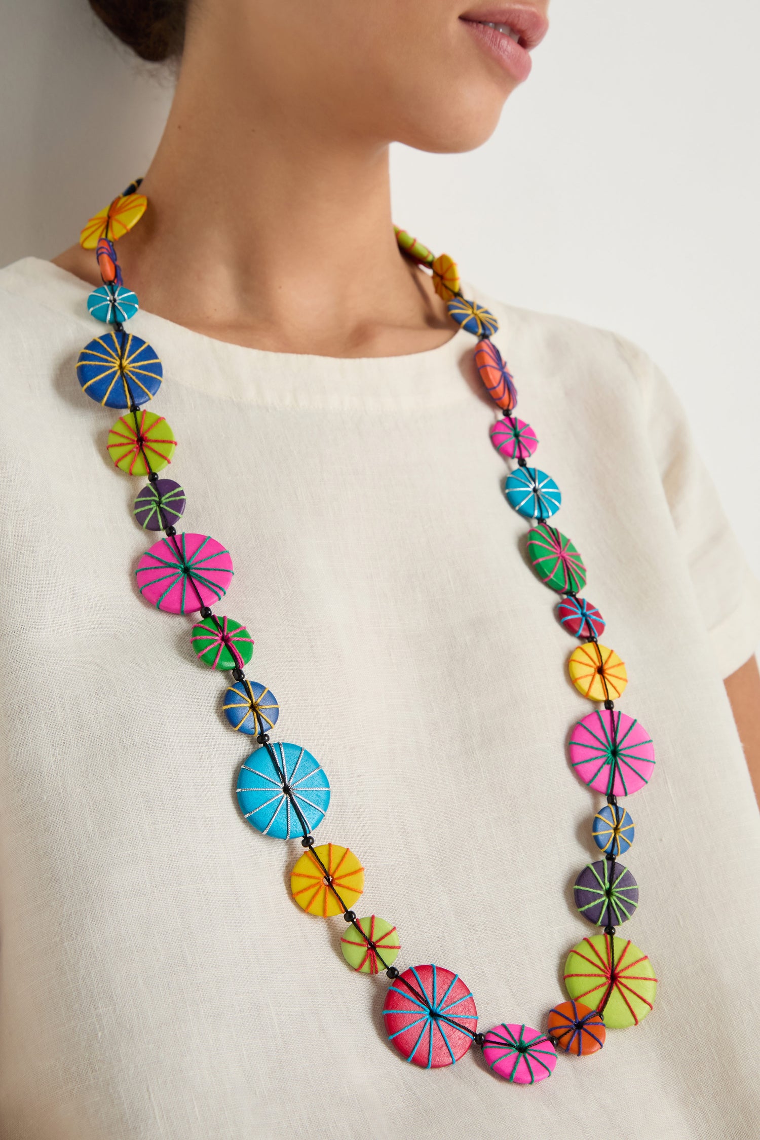 A woman wearing a white top and the Long Wooden Disc Necklace, featuring colorful round beads with geometric stitching patterns and subtle thread wrap details.