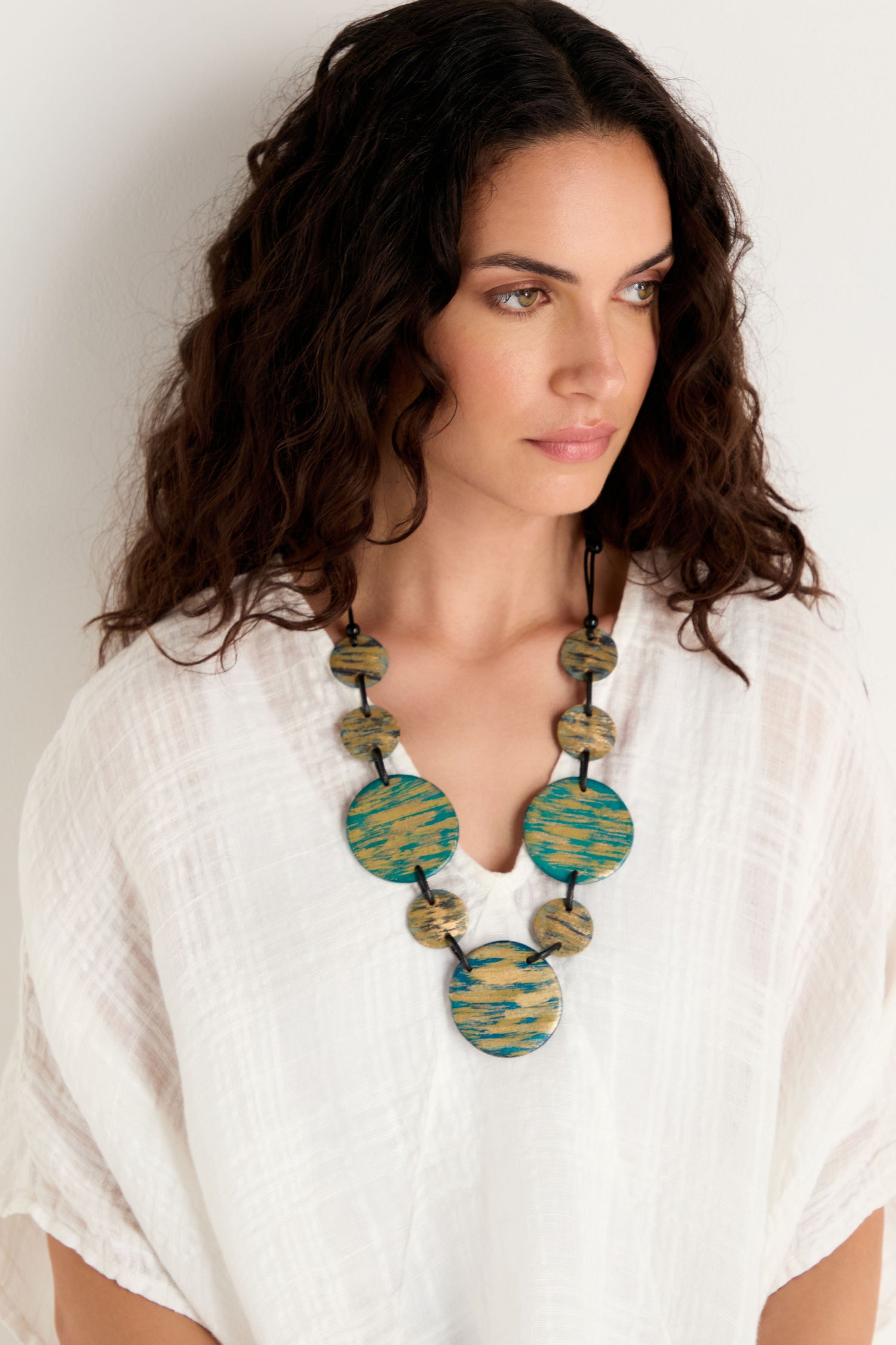 Woman with wavy dark hair wears a white textured blouse and the Painted Disc Necklace, featuring large blue and green circular accents, standing against a plain white background.