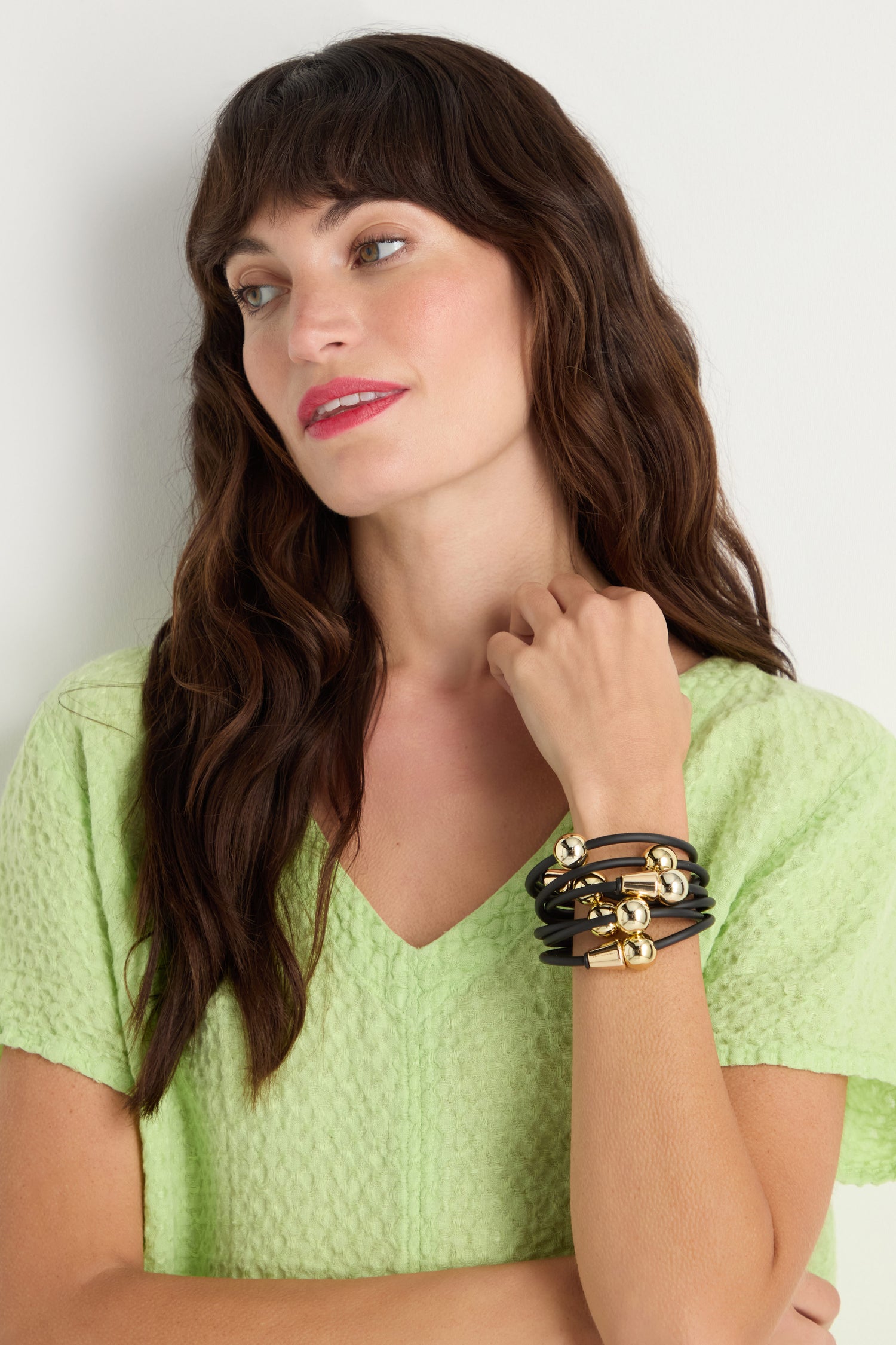 A woman with wavy brown hair in a textured light green top looks to the side, showcasing a stack of black and gold bracelets on her wrist, featuring the Metal Spheres Bracelet—a striking, sculptural design handmade in France.