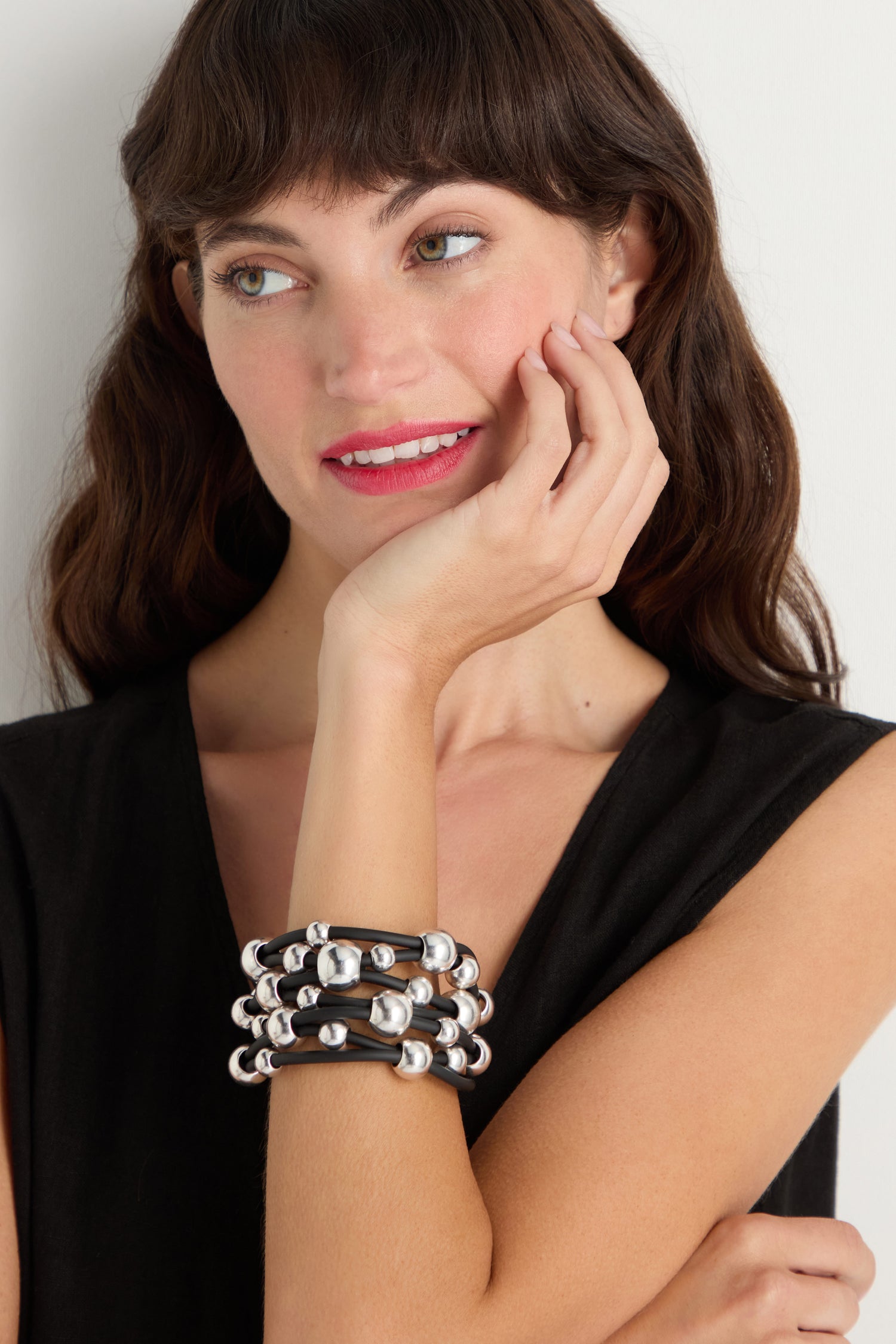 A woman with wavy brown hair wears a sleeveless black top and the Metal Spheres Bracelet—a chunky, sculptural black bracelet featuring metal spheres—resting her hand on her cheek while looking to the side.