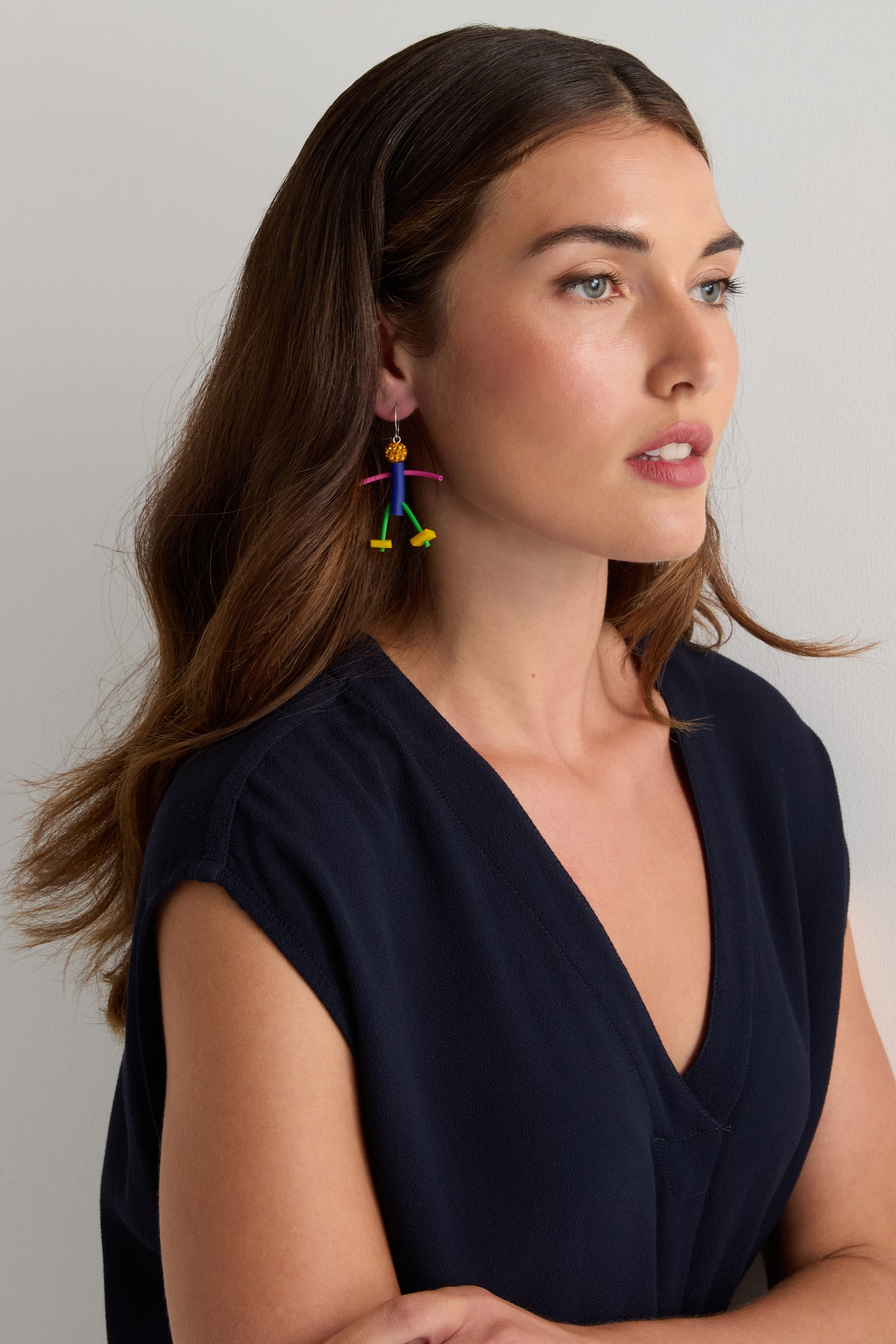 Woman with long brown hair wears a navy V-neck top and Beautiful People Earrings by Samuel Coraux, featuring playful abstract stick figures, while posing against a plain light background.