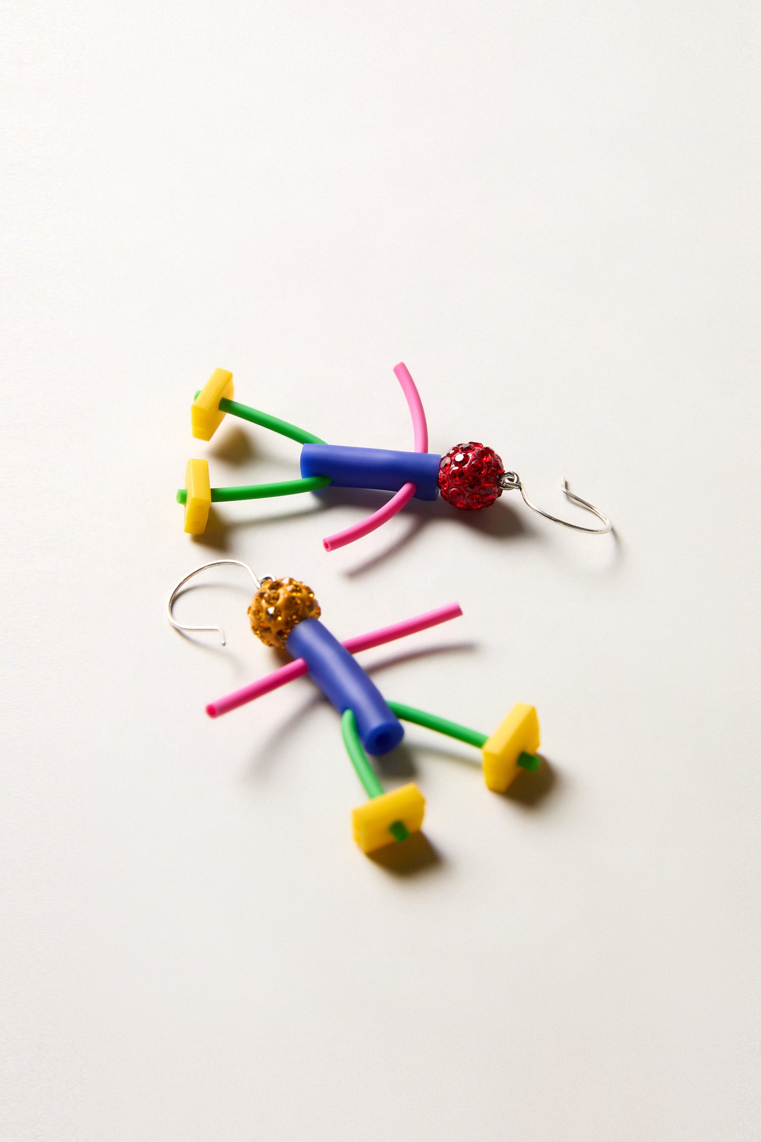 The Beautiful People Earrings by Samuel Coraux feature abstract stick figure designs with blue bodies, pink arms, yellow feet, and textured round heads, displayed on a white surface.