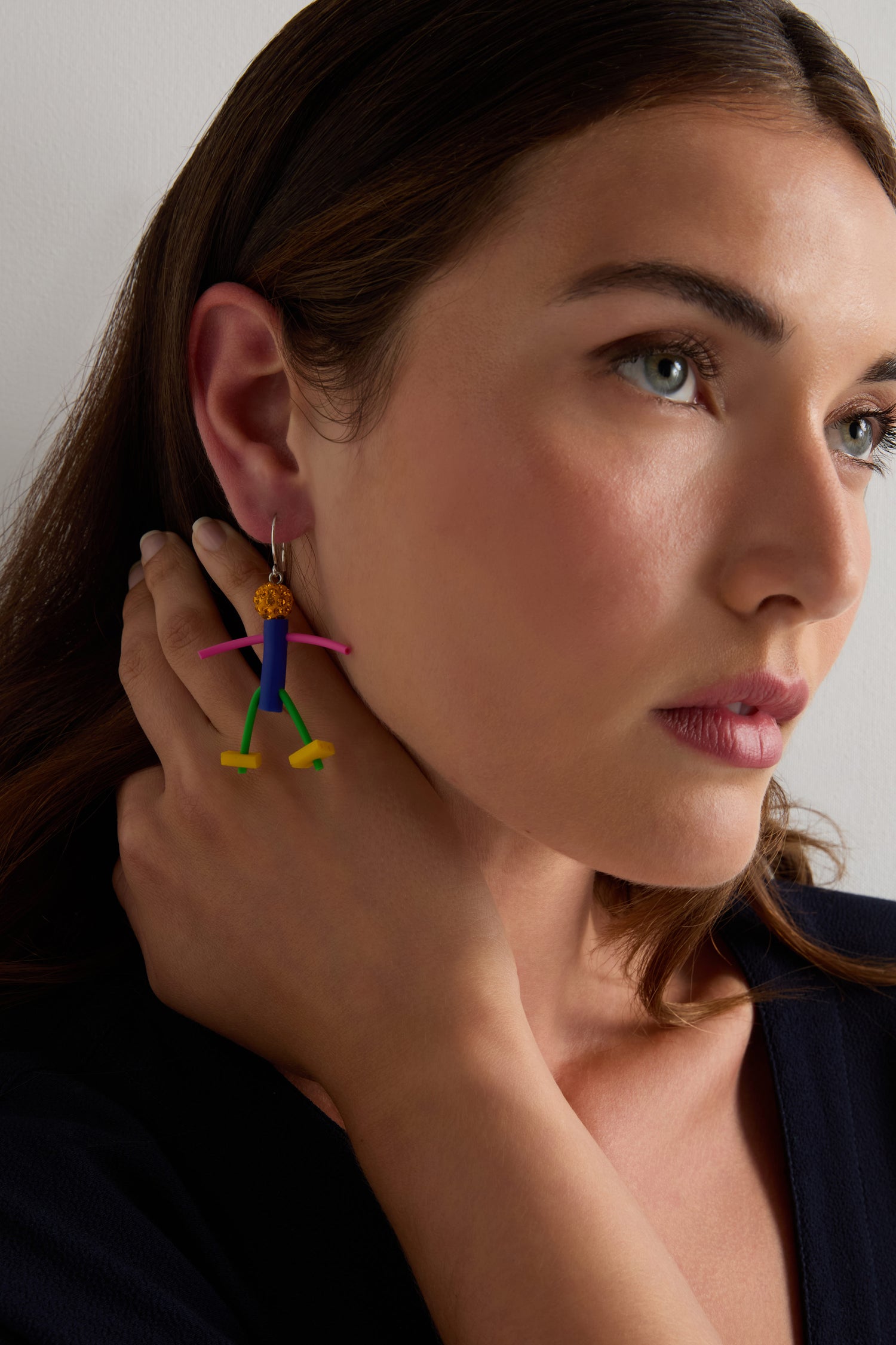A woman with brown hair touches her neck, wearing playful Beautiful People Earrings and a dark top, against a plain background.