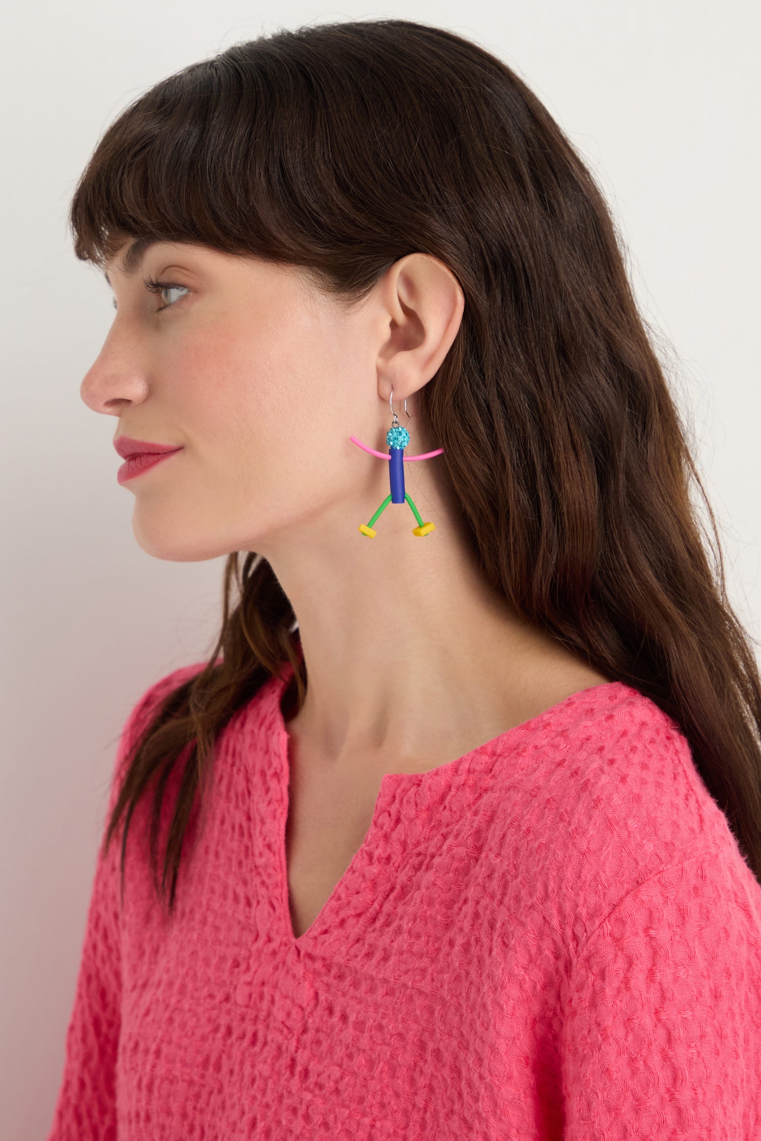 Woman with long brown hair in a textured pink top wears Beautiful People Earrings—colorful, abstract figure earrings handmade in Paris—shown in profile against a plain background.
