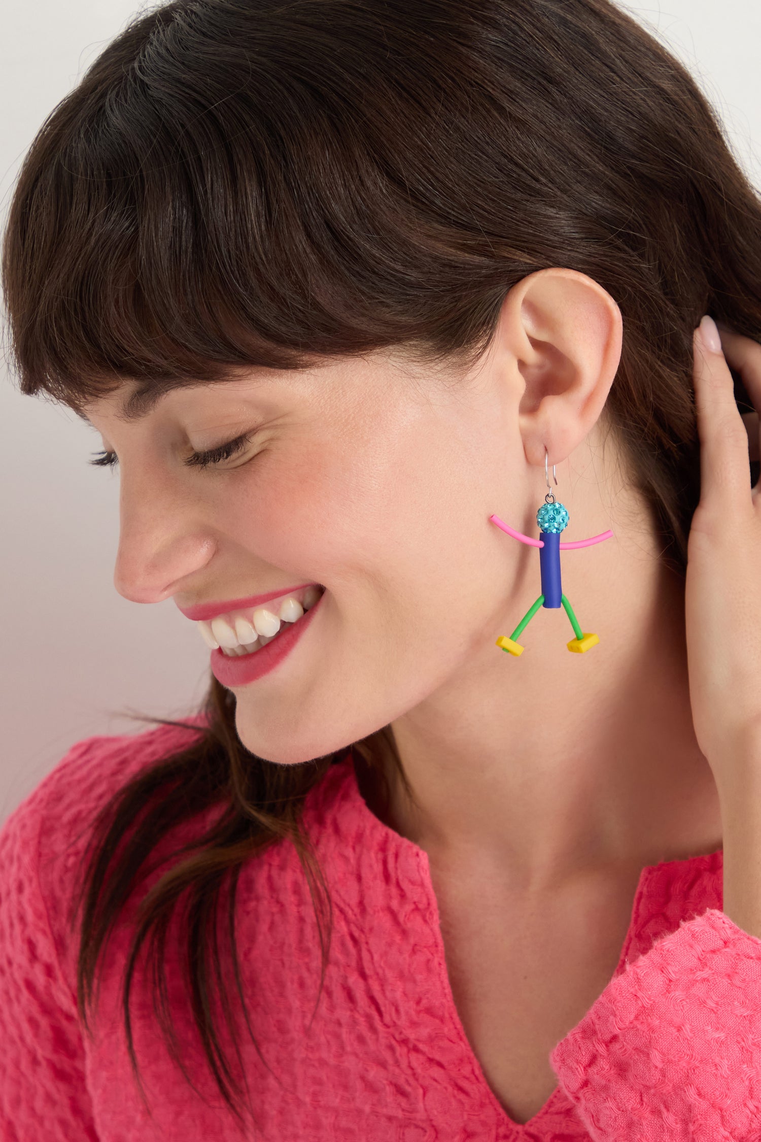 A woman with brown hair smiles, wearing a bright pink textured top and the Beautiful People Earrings—colorful, abstract, handmade in Paris—for a charming, playful touch while she holds a hand near her ear.