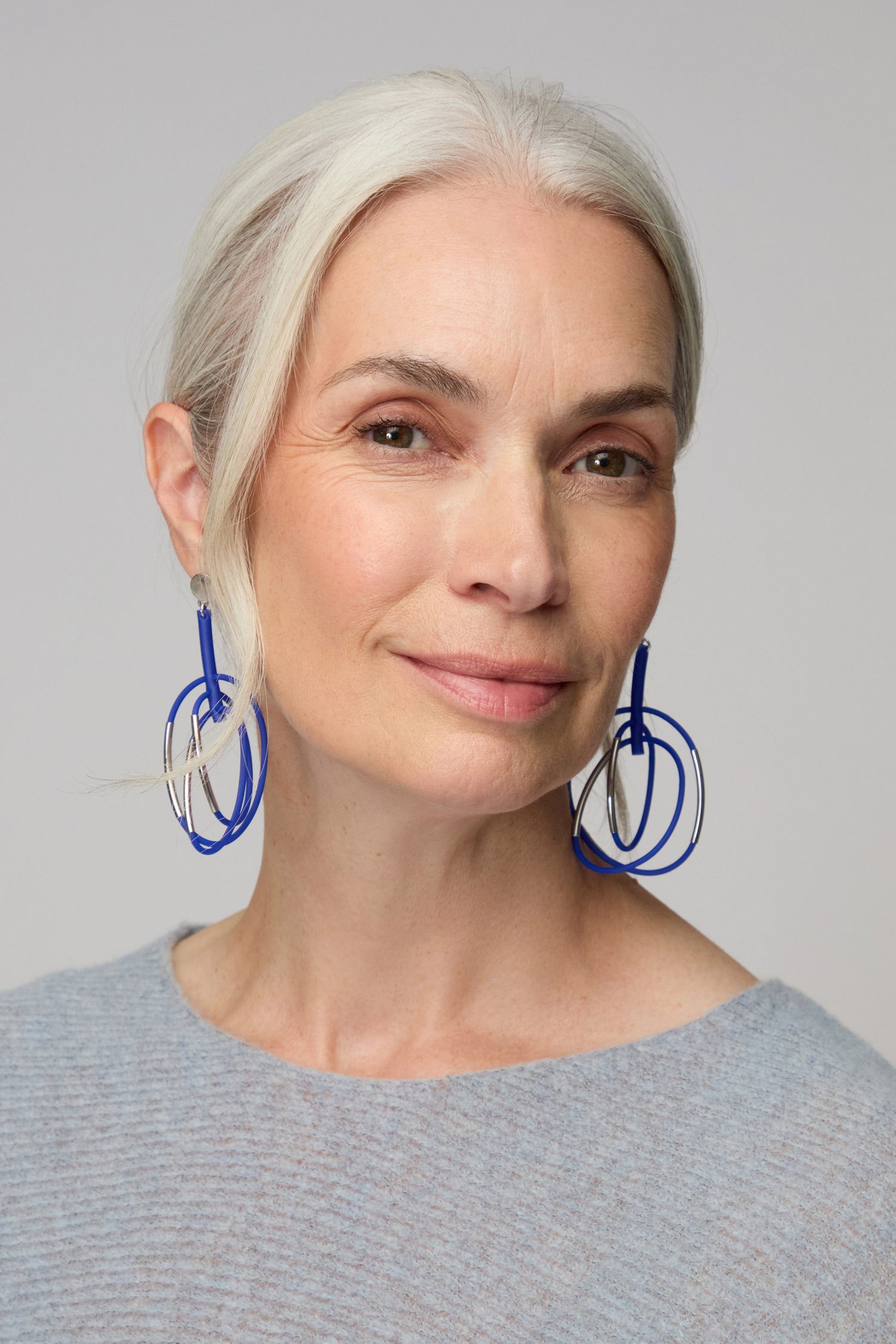 A woman with gray hair wears Carousel Earrings and a light gray top, looking at the camera with a slight smile against a neutral background.