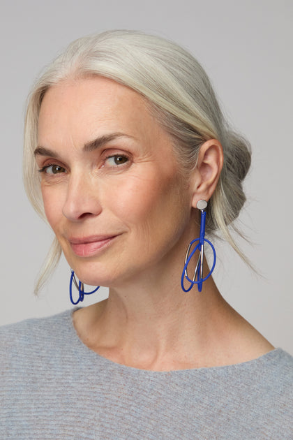 With her gray hair in a low bun and dressed in a light blue top, she poses against a plain background, highlighting her bold style with the Carousel Earrings.