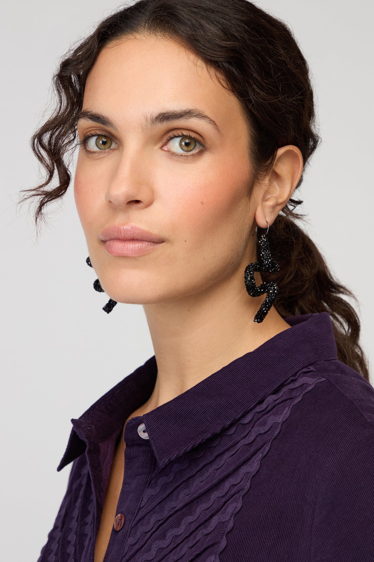 A woman with wavy brown hair in a ponytail, wearing a purple collared shirt and bold Disco Sketch Black Earrings, looks at the camera with a neutral expression.