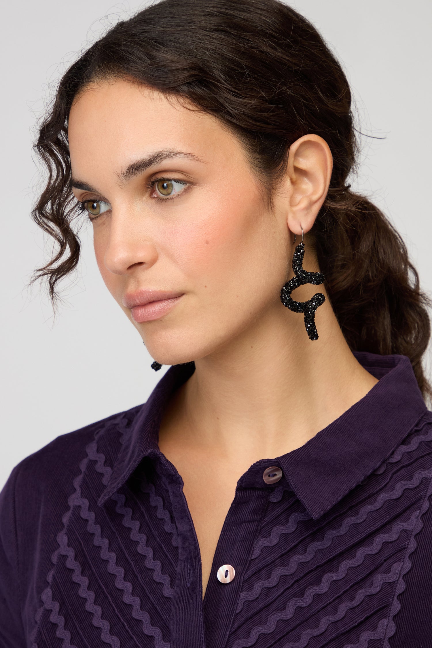 A woman with curly brown hair in a ponytail wears a purple textured shirt and statement jewellery, including Disco Sketch Black Earrings, as she looks slightly to the side against a plain background.