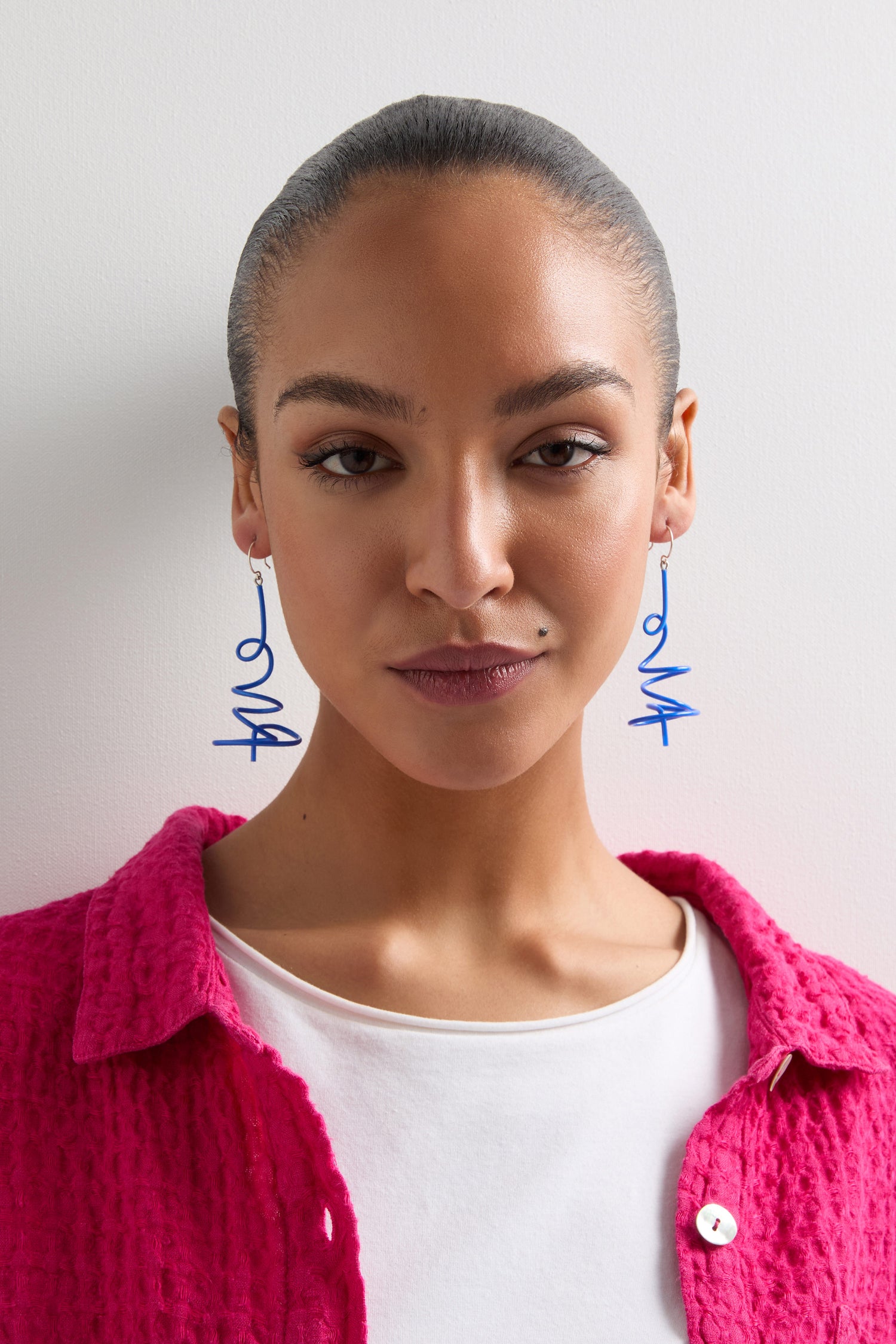 A person with pulled-back hair confidently showcases Squiggle Earrings and a textured pink jacket over a white top against a white background, perfectly embracing bold color schemes to highlight their unique style.