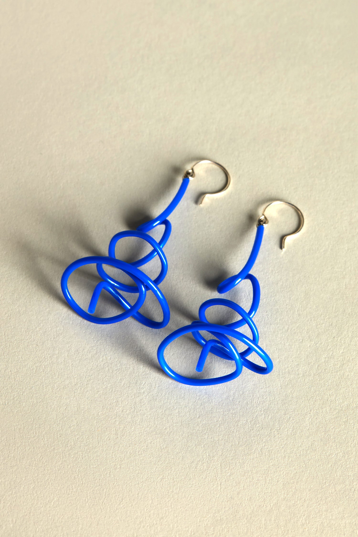 The Squiggle Earrings, featuring blue abstract shapes with silver hooks and echoing Samuel Coraux's bold color schemes, rest on a light surface.