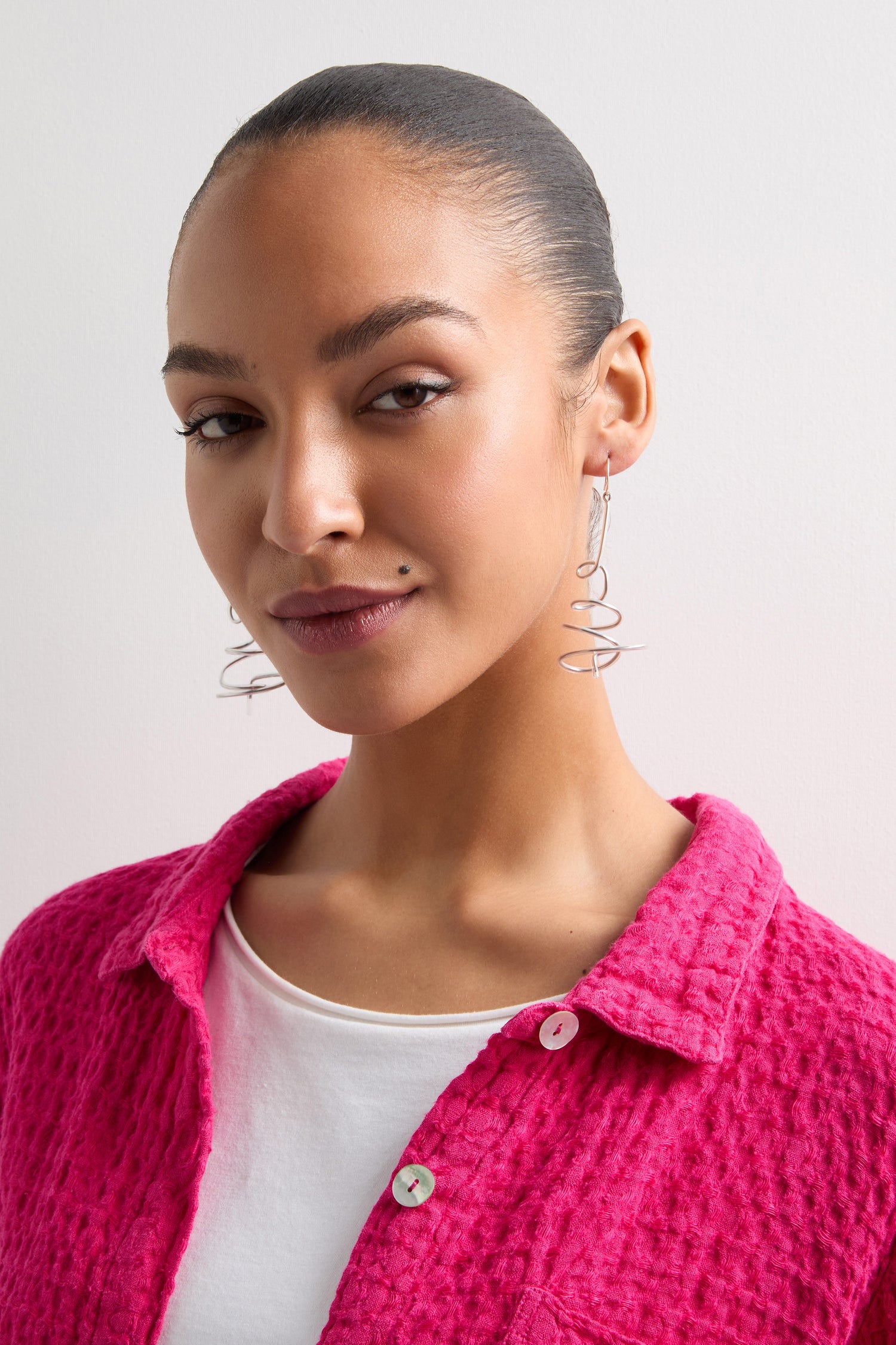 Wearing a pink textured shirt and white top, paired with bold Squiggle Earrings reminiscent of Samuel Coraux's designs, a person confidently looks at the camera.