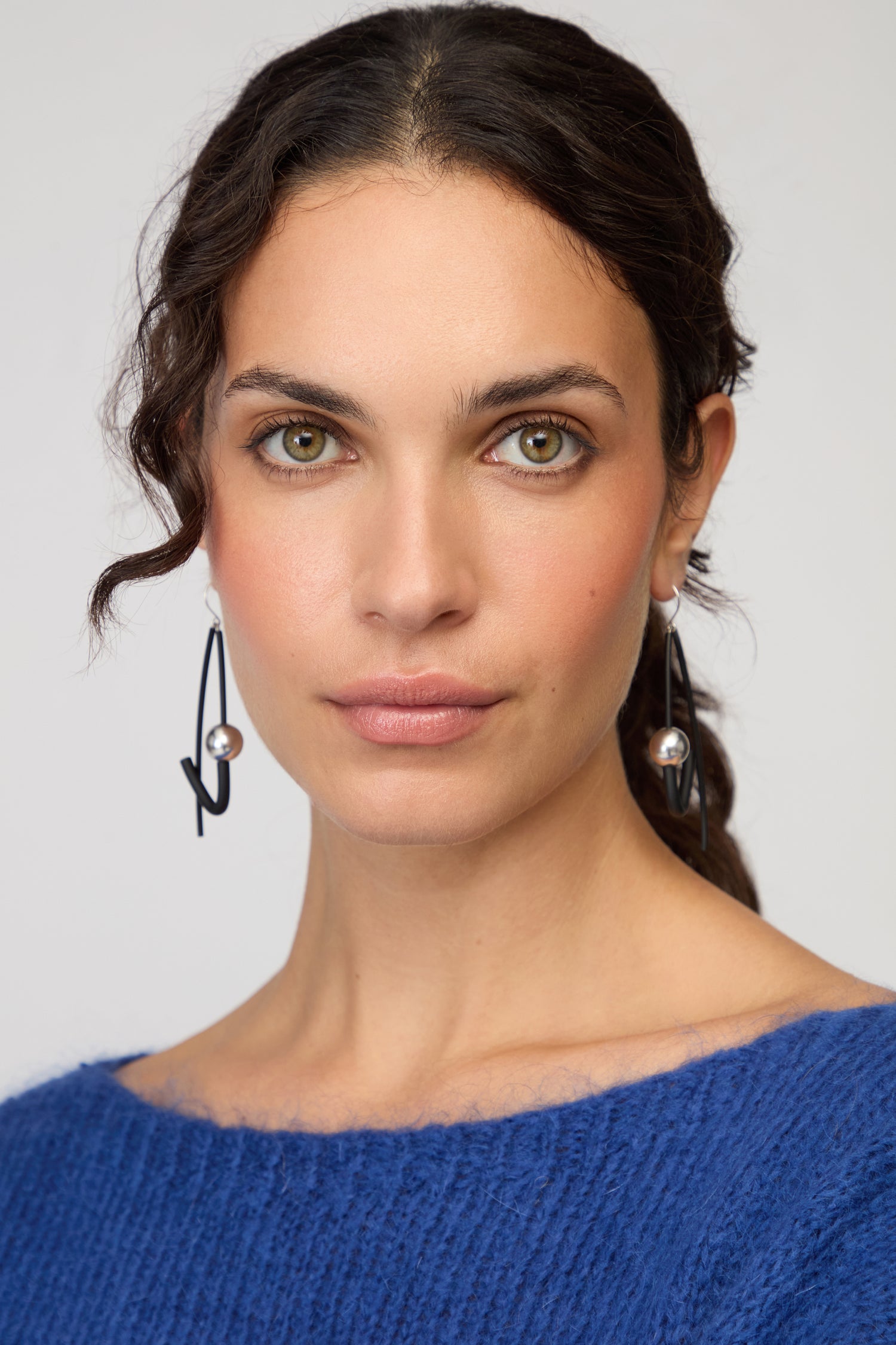 Woman with wavy brown hair, wearing a blue sweater and eye-catching Silver Sphere Earrings by Samuel Coraux, looks directly at the camera against a plain background.