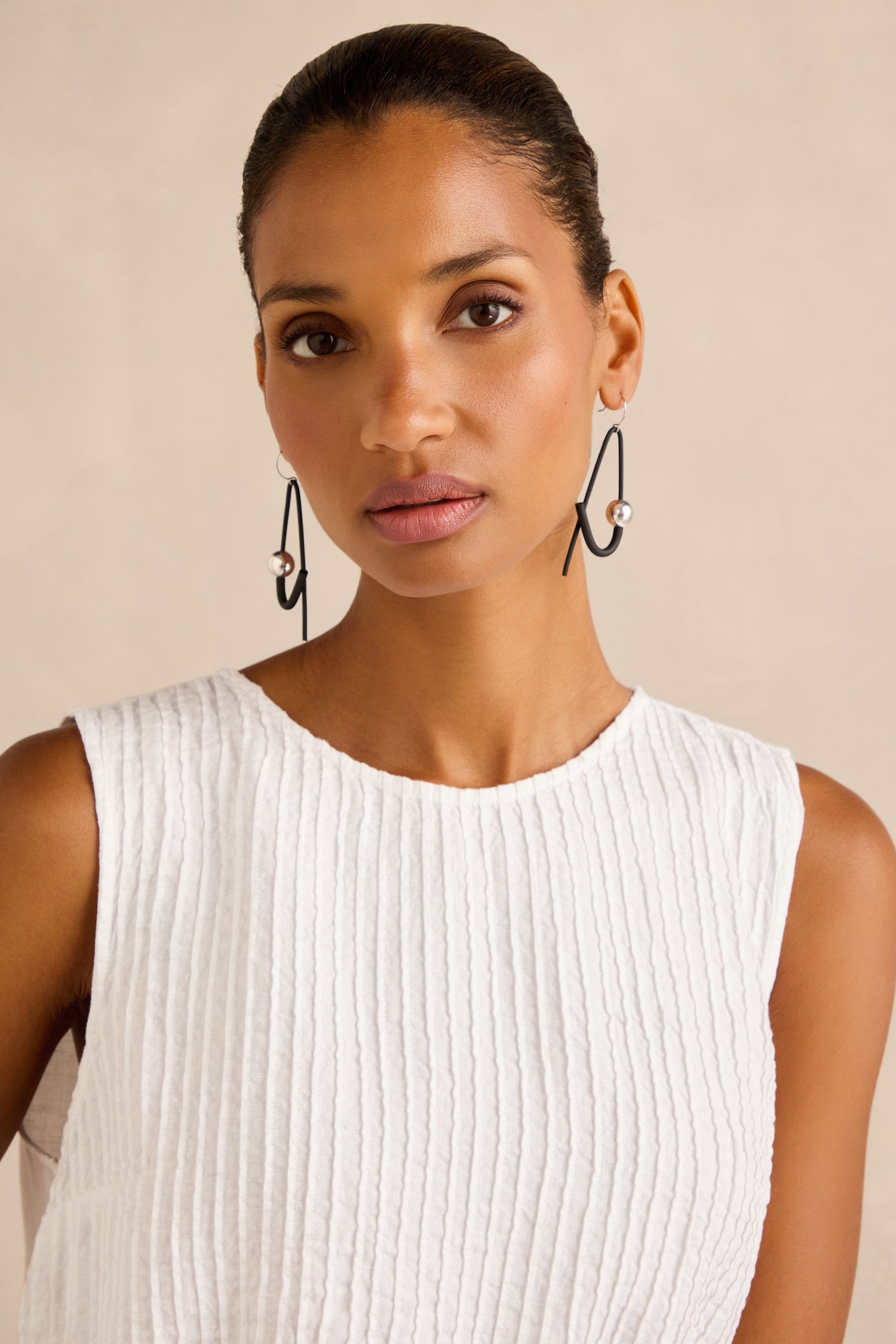 A woman wearing a sleeveless white textured top and large, contemporary Sphere Earrings—handmade in Paris—stands against a plain, light background.