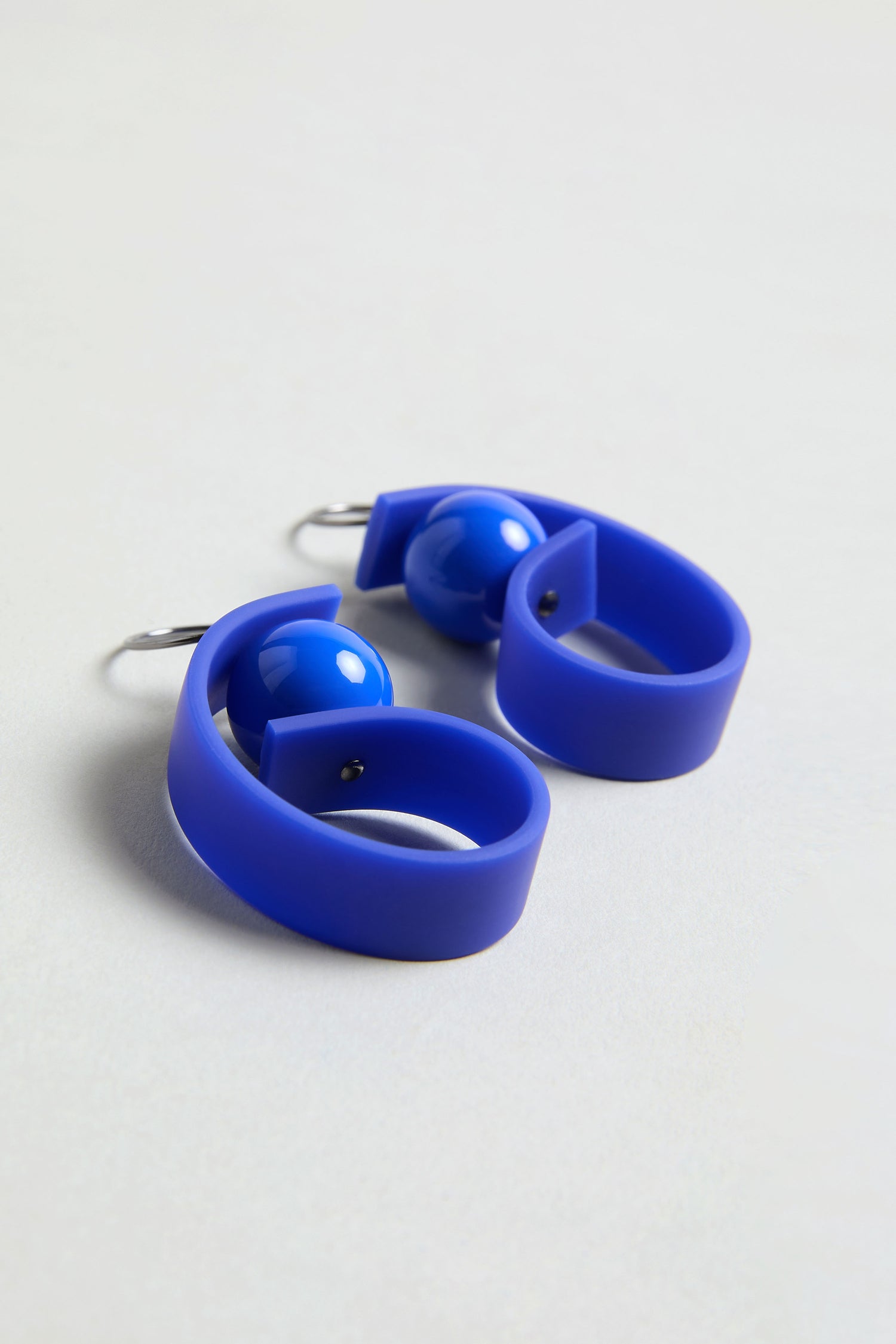 The Penseur Earrings are modern royal blue statement earrings with a curved, looped design and glossy blue bead accents—perfect for lovers of sculptural jewellery.