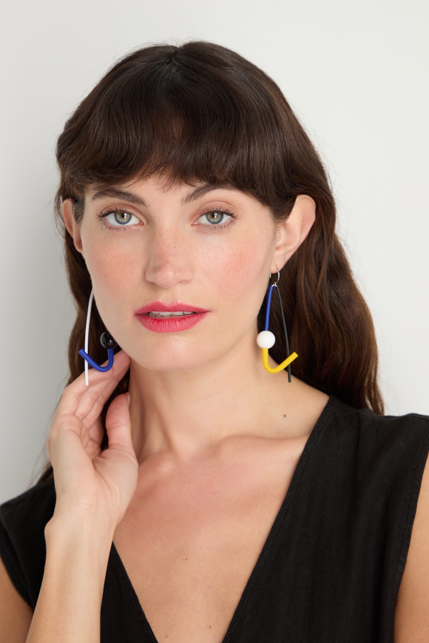A woman with long brown hair and a black sleeveless top models the handmade Kandinsky Sphere Earrings. She gazes at the camera, hand lightly touching one of the geometric, colorful earrings crafted in Paris.
