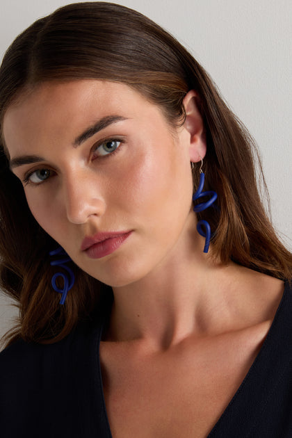 A woman with brown hair wears Sketch Earrings by Samuel Coraux—lightweight rubber with a sculptural spiral design—and a dark top, looking slightly to the side against a light background.