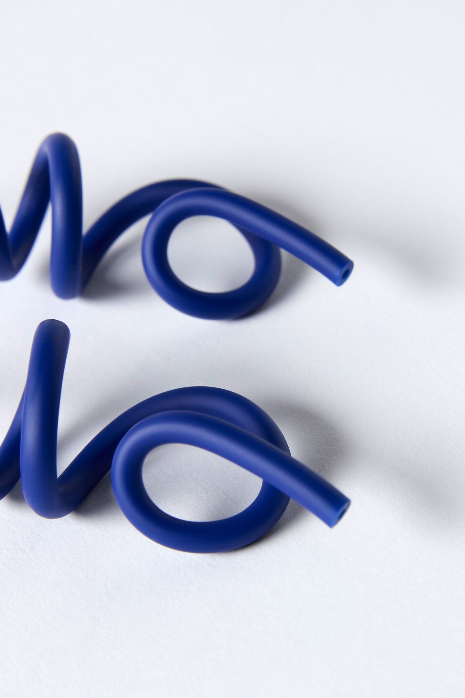 Two blue plastic Sketch Earrings with playful loops rest parallel on a white surface, their sculptural curves reminiscent of Samuel Coraux's spiral designs.