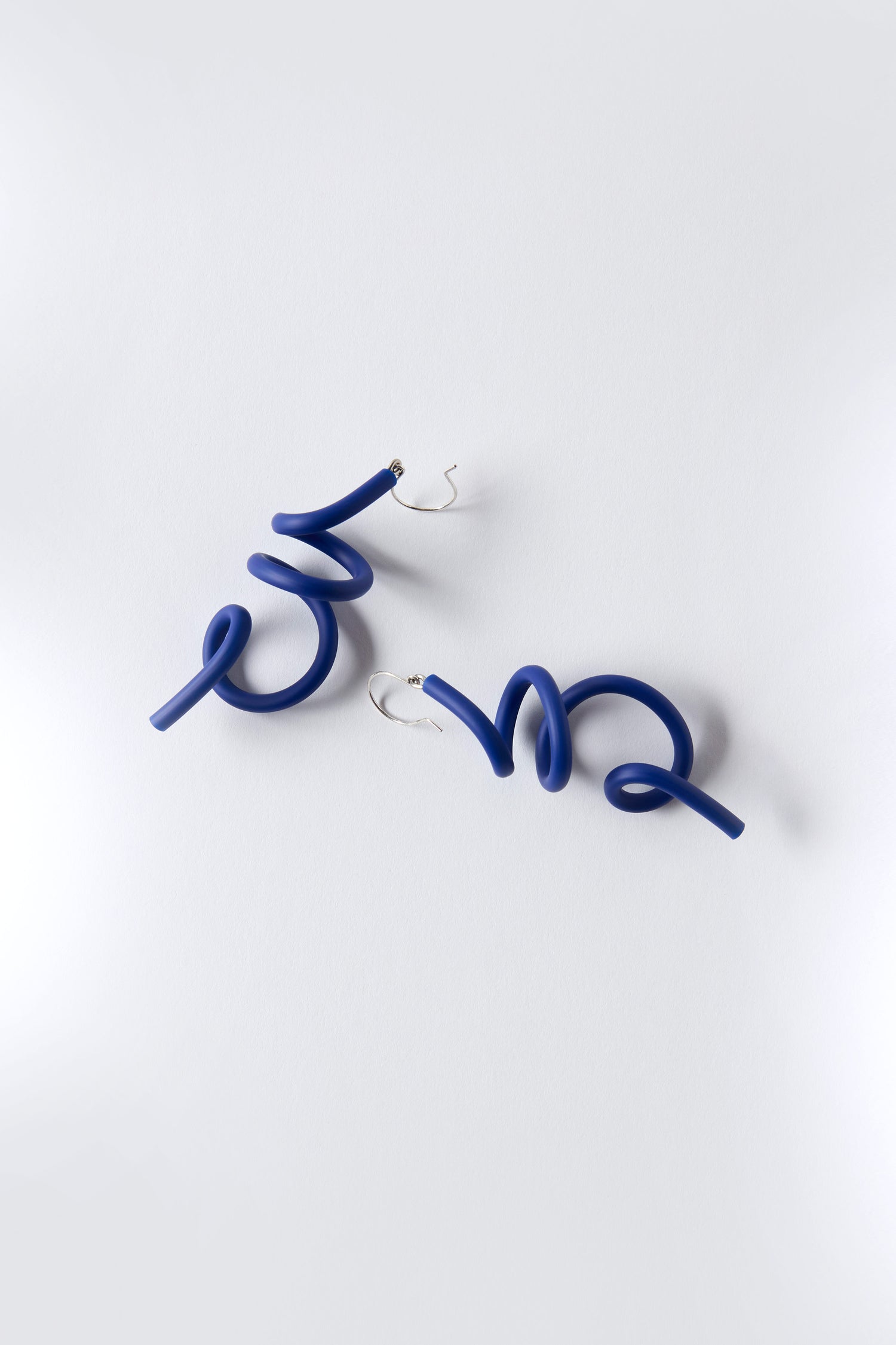 The Sketch Earrings by Samuel Coraux are blue sculptural spiral earrings crafted from lightweight rubber with silver hooks, shown on a plain white background.