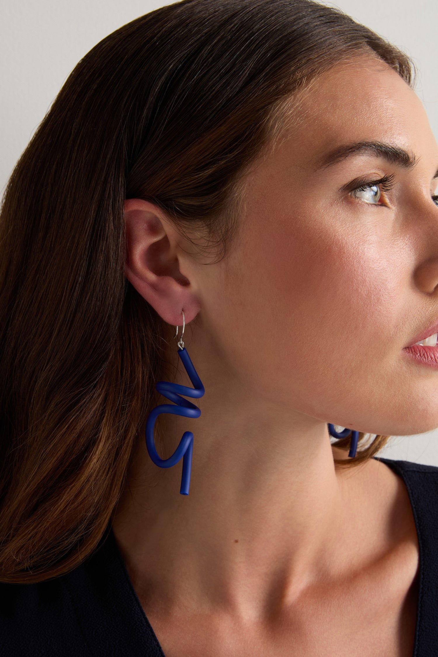 A woman with brown hair models the blue Sketch Earrings by Samuel Coraux, paired with a dark top, in a close-up side profile.