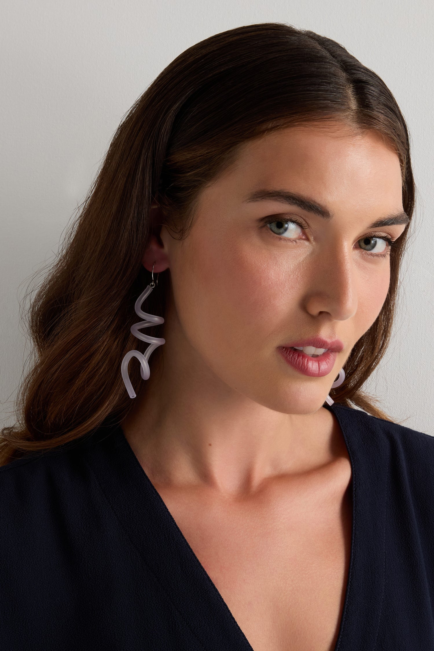 Woman with wavy brown hair wears a navy top and Sketch Earrings by Samuel Coraux—lightweight rubber earrings featuring large, abstract lavender shapes—while standing against a plain light background.