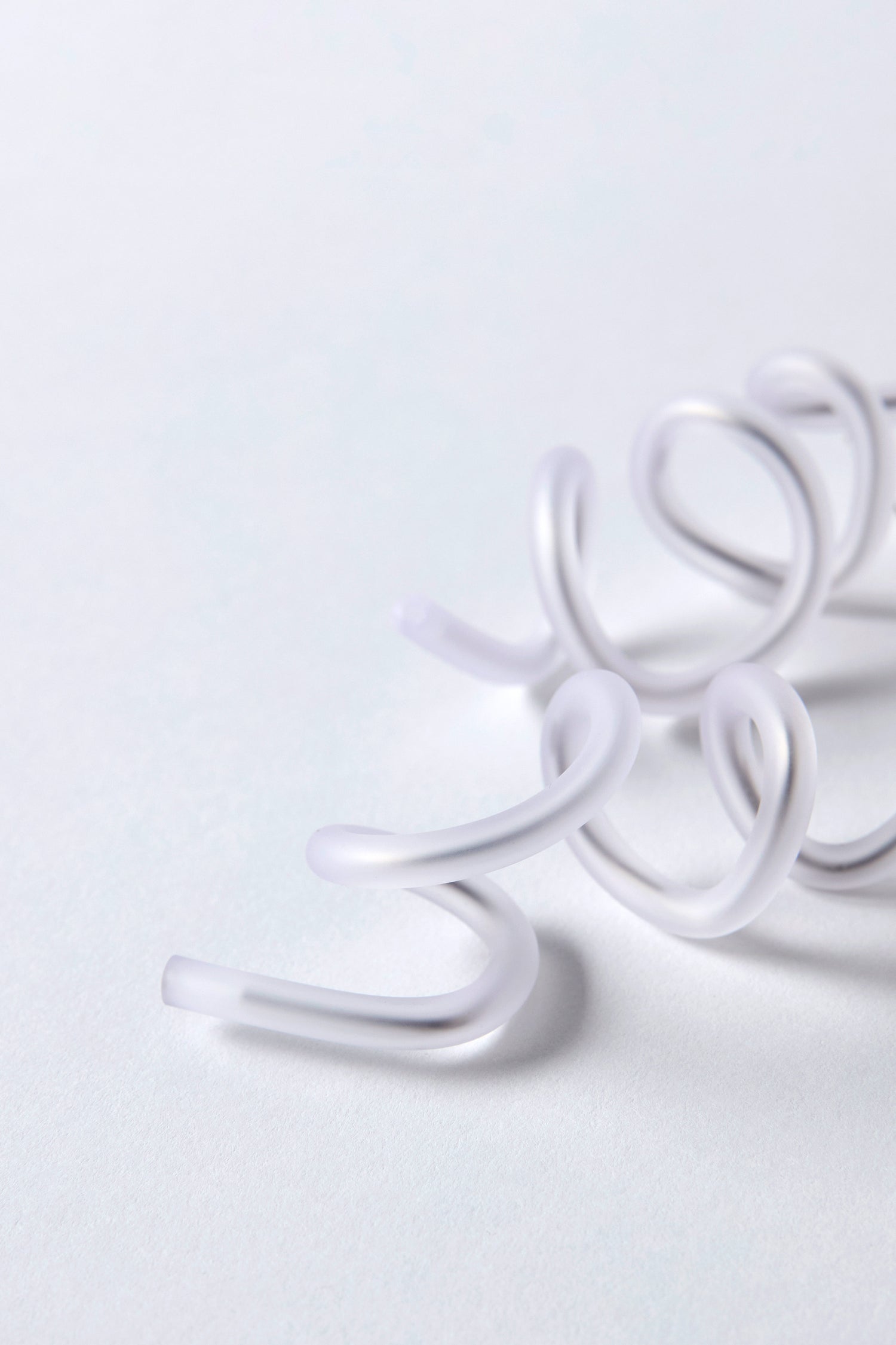 Close-up of Sketch Earrings, translucent and coiled in a sculptural spiral, displayed on a light gray surface.