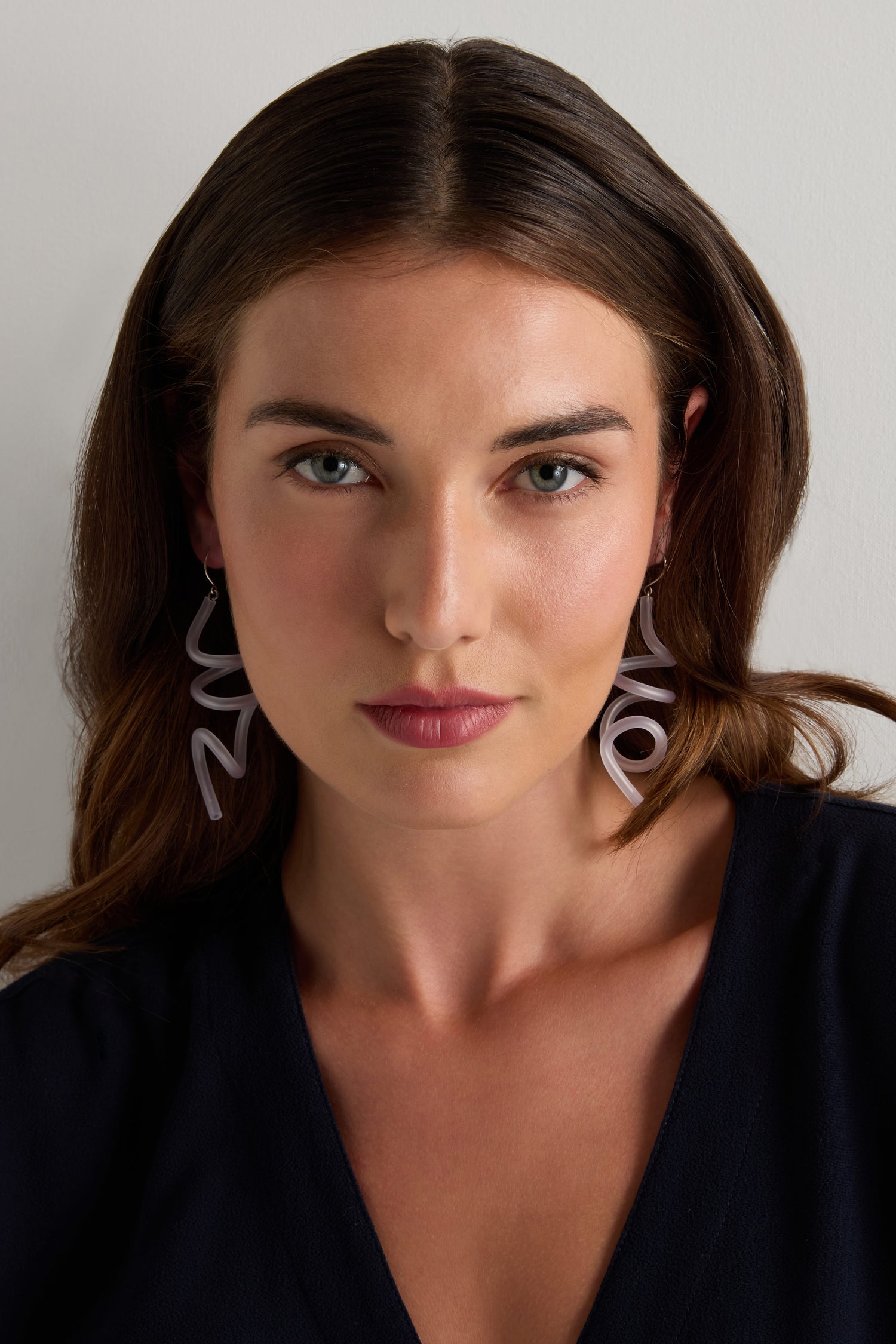 Woman with shoulder-length brown hair wearing Sketch Earrings by Samuel Coraux and a dark V-neck top, facing forward against a plain light background.