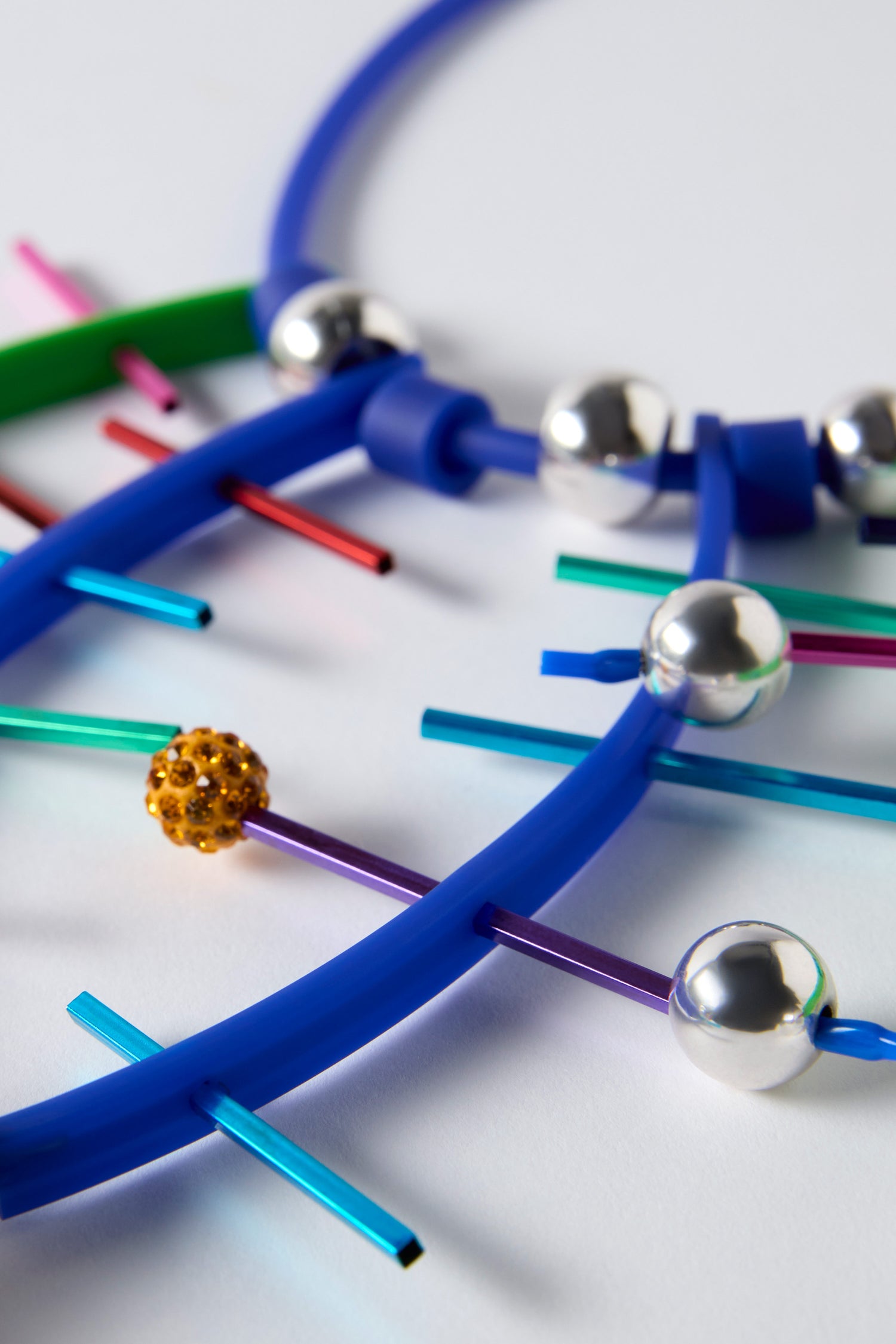 Close-up of the Fame Necklace, handmade in Paris by Samuel Coraux, showcasing blue and green curved tubes, metallic spheres, colored rods, and a single gold bead against a white background.